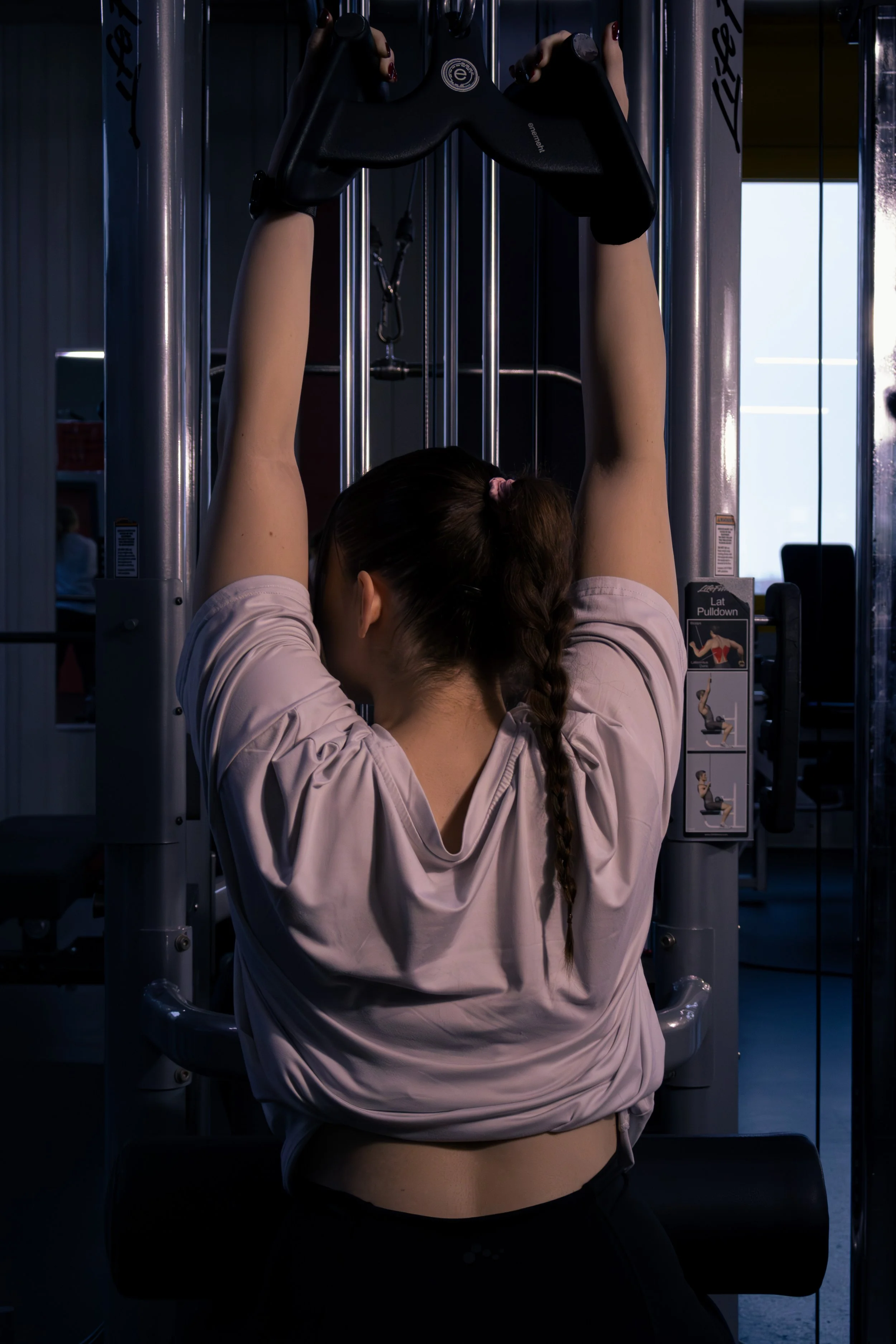 Eine Frau mit braunem Haar in einem Zopf, die an einem mittelgroßen Fitnessgerät hängt und die Arme nach oben streckt, trägt eine rosa Sportjacke, in einem dunklen Trainingsraum.
