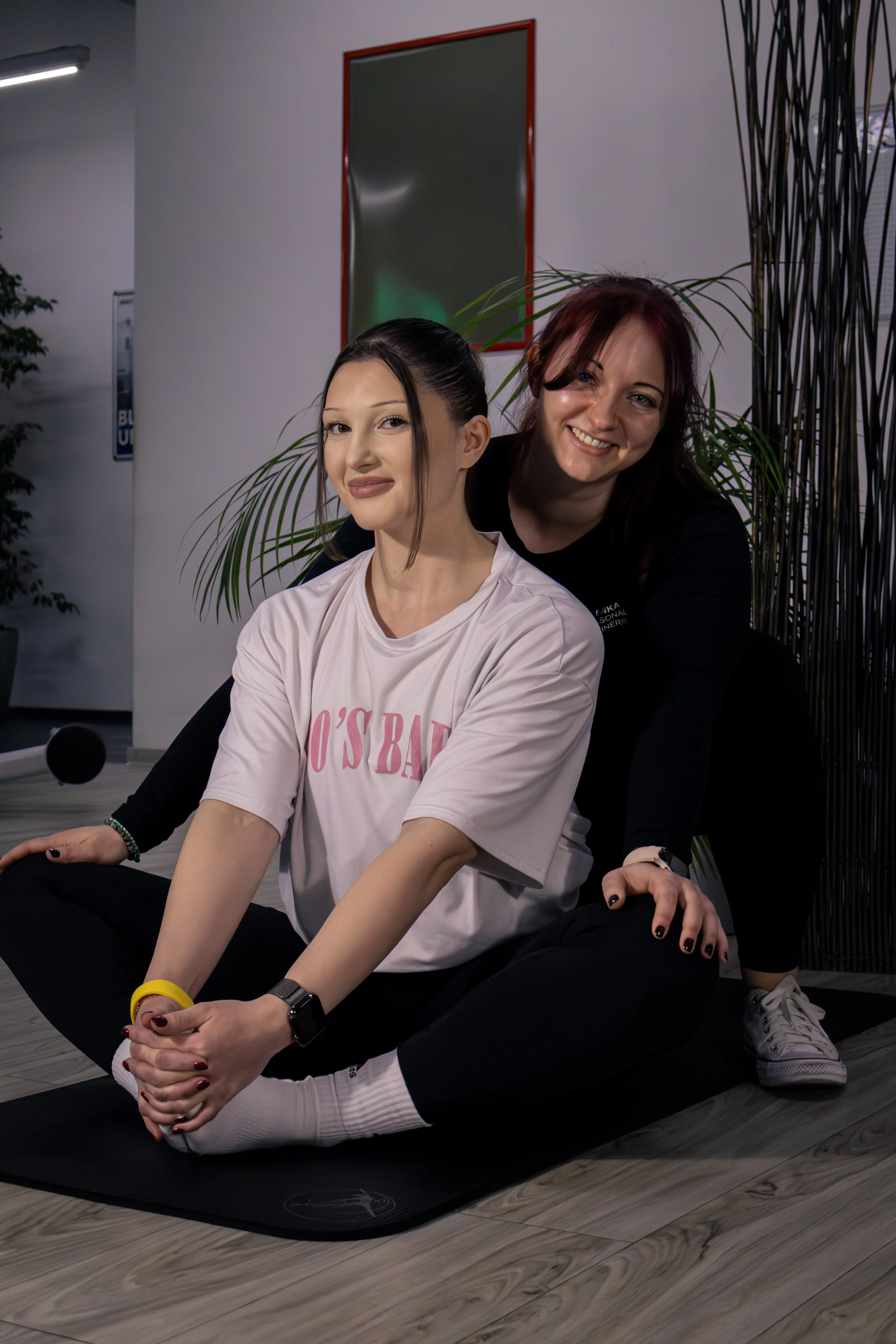 Zwei Frauen in einer Fitnessstudio-Umgebung, eine sitzt auf dem Boden mit verschränkten Beinen und der anderen steht dahinter, lächelnd. Beide tragen sportliche Kleidung und sind auf Yogamatten, mit Pflanzen und Spiegeln im Hintergrund.