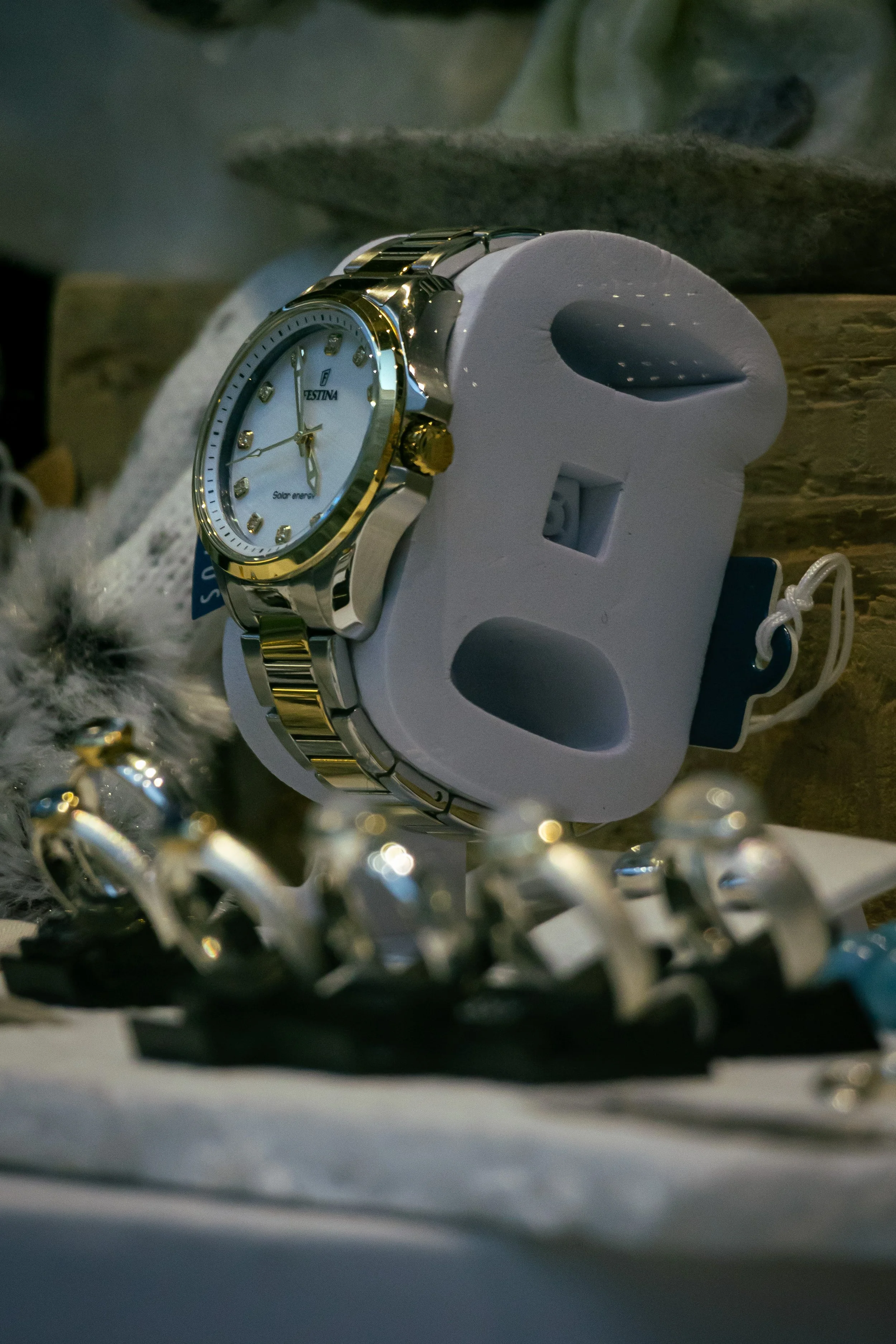 Ein goldener Armbanduhr mit weißen Zifferblatt und Diamantenmarkierungen auf einem weißen Uhrenständer. Im Hintergrund sind Ketten und Schmuckstücke sichtbar.