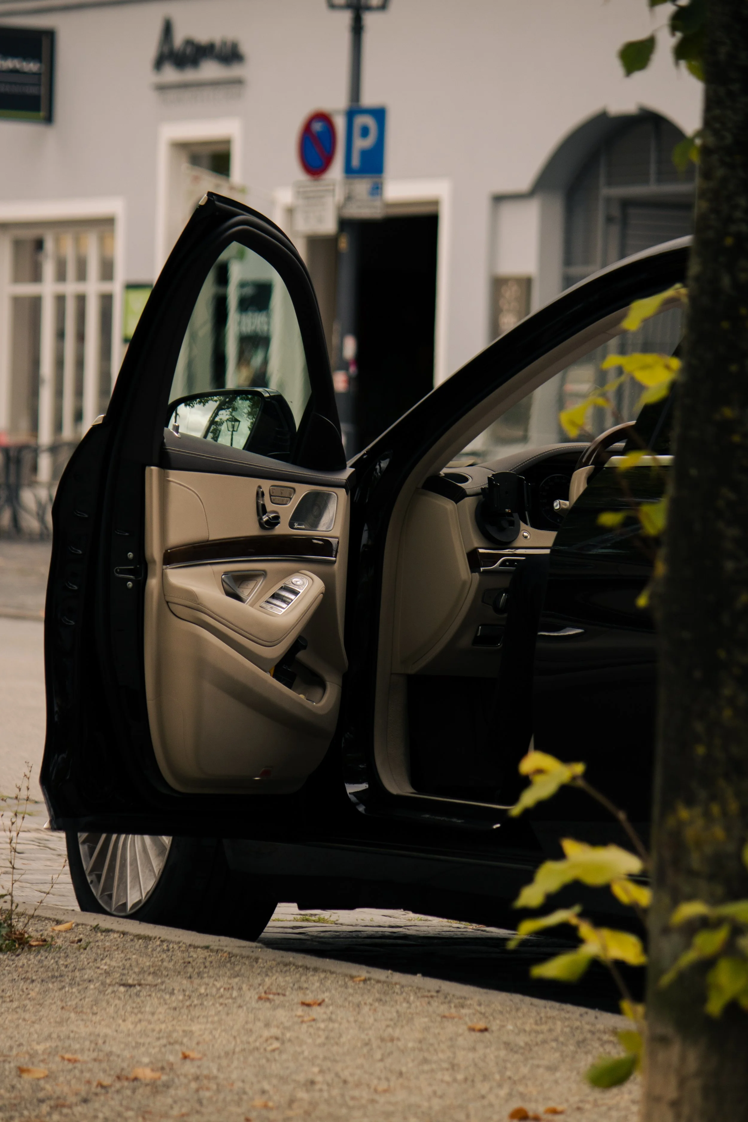Das Innere eines schwarzen Autos mit offenem Fahrertür, Blick auf die Instrumententafel. Im Hintergrund eine weiße Fassade mit Park- und Halteverbotsschilder, es sind einige Blätter im Vordergrund sichtbar.