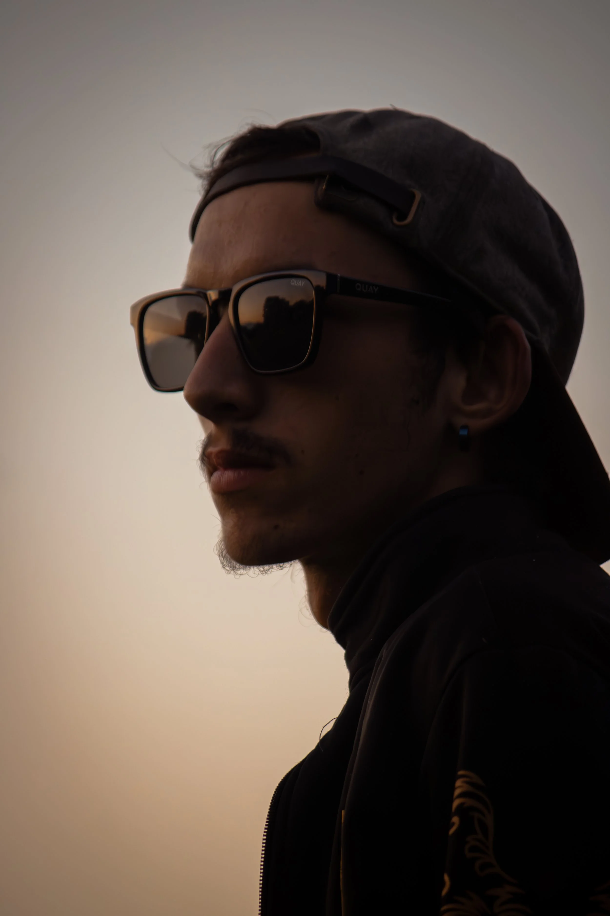 Nahaufnahme eines Mannes mit Halbprofil, Sonnenbrille und Kapuzenshirt vor hellem Himmel bei Sonnenuntergang.