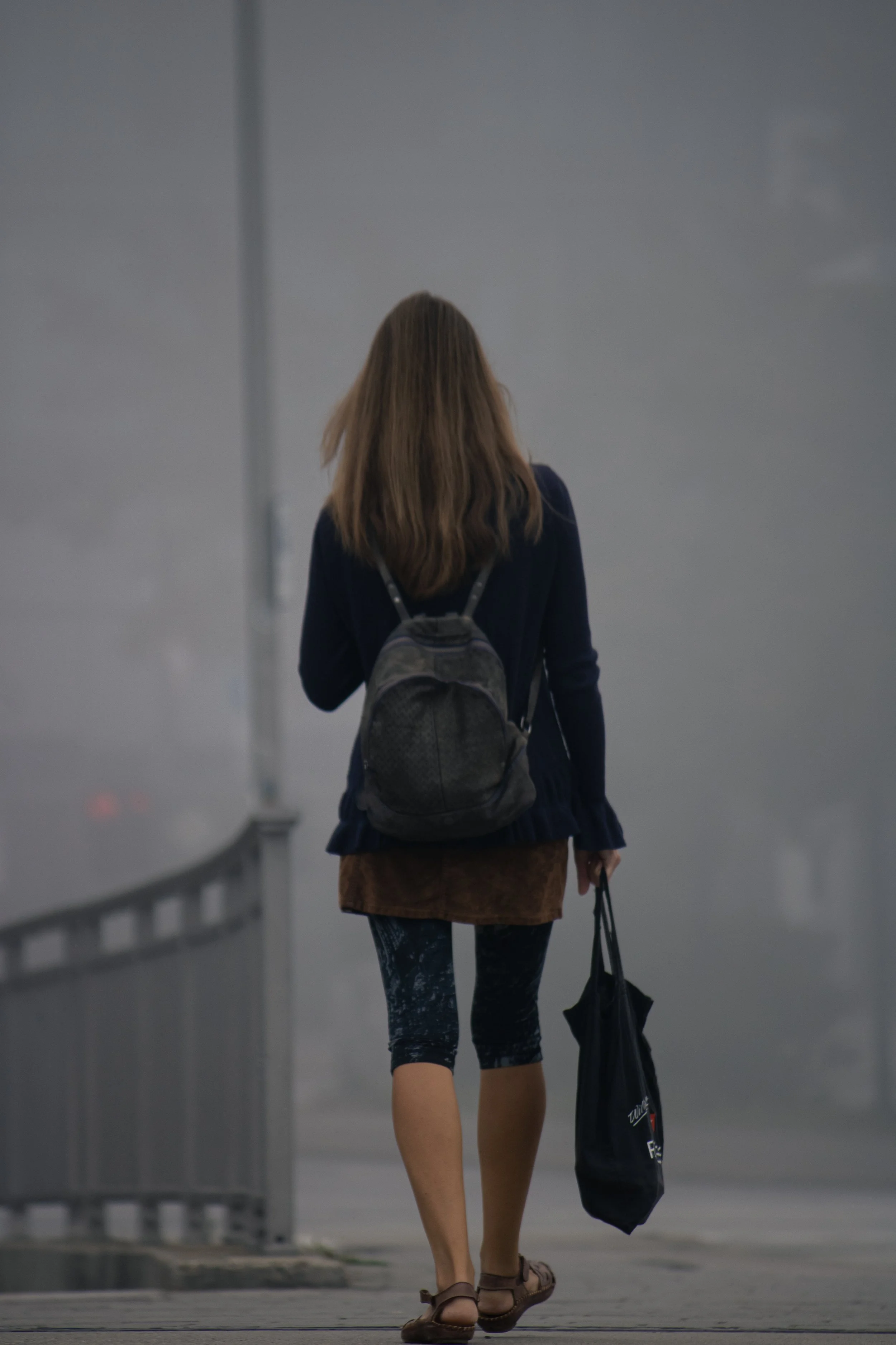 Eine Person mit braunen Haaren, die einen Rucksack trägt und einen schwarzen Beutel hält, geht an einer Brücke in einer urbanen Umgebung.