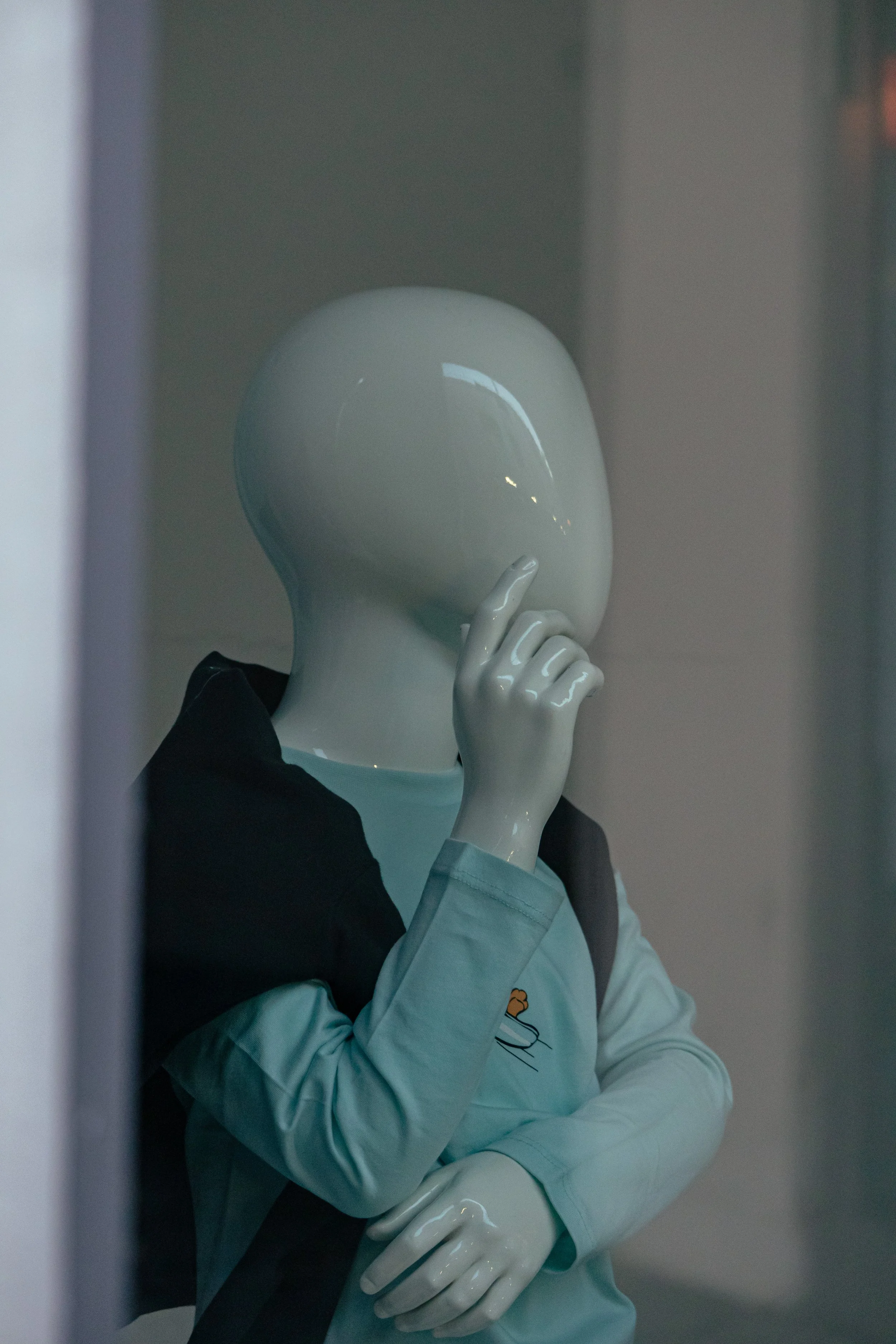 Skulptur einer Figur mit einem weißen, runden Kopf, die mit einer Hand an den Lippen hält, in einem Geschäftsfenster. Die Figur trägt eine hellblaue Jacke mit einem Logo auf der Tasche.