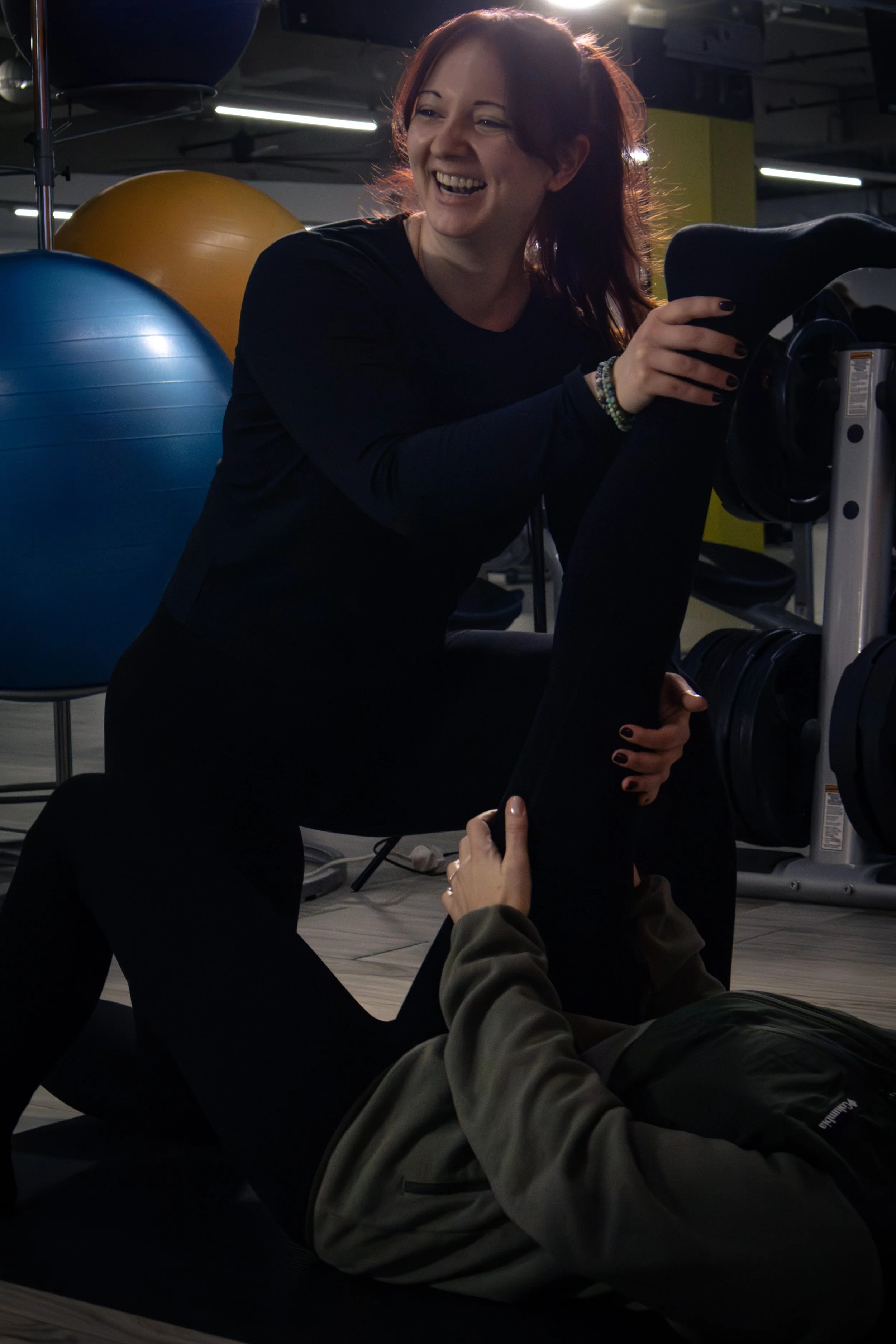 Zwei Frauen im Fitnessstudio, eine steht lachend und eine liegt auf dem Boden, während sie sich gegenseitig unterstützen, um Klimmzüge zu machen. Es gibt Gymnastikbälle im Hintergrund.