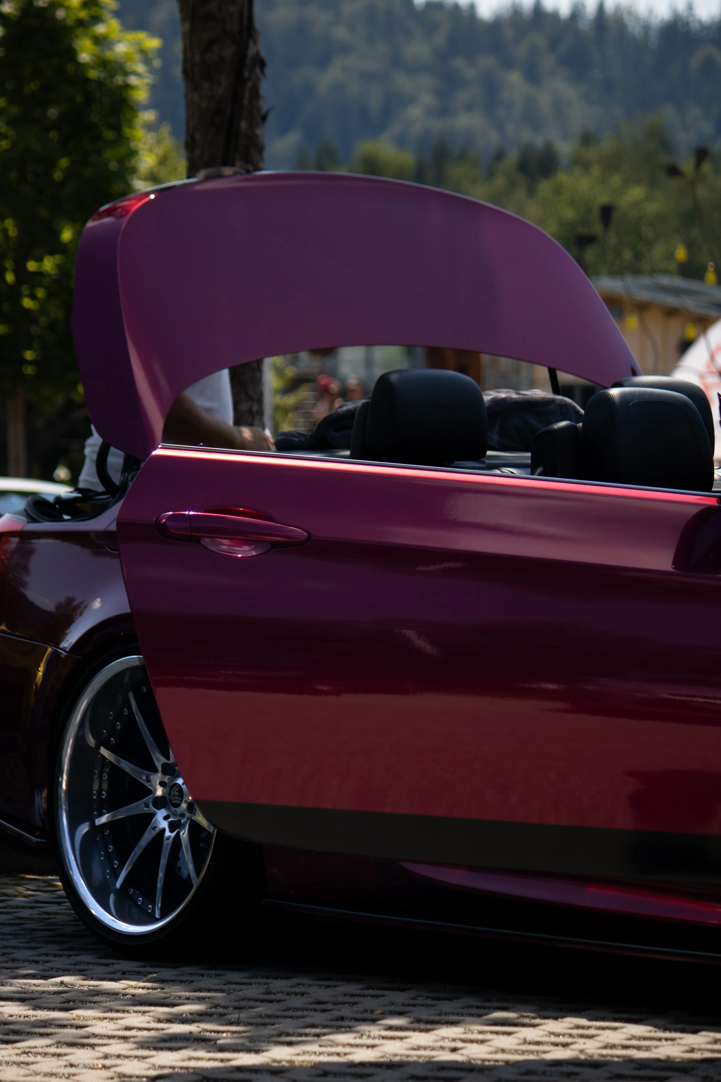 Ein rotes Convertible-Auto mit offenem Dach auf einem gepflasterten Straßenabschnitt im Freien, im Hintergrund Bäume und eine bergige Landschaft.