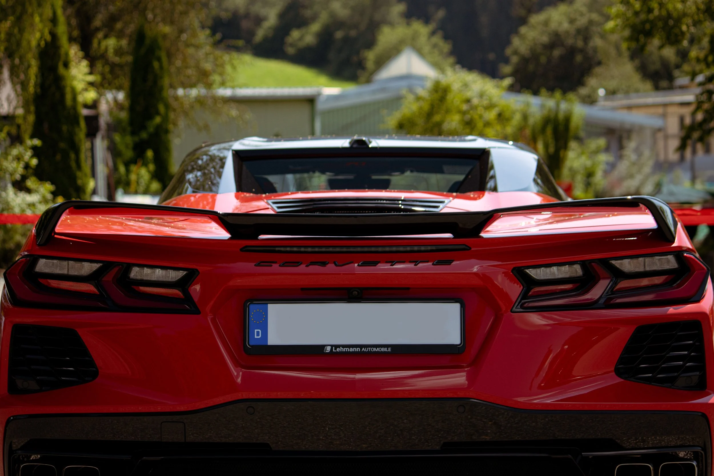 Roter Sportwagen von hinten, in einer Outdoor-Umgebung mit Bäumen und Gebäuden im Hintergrund.