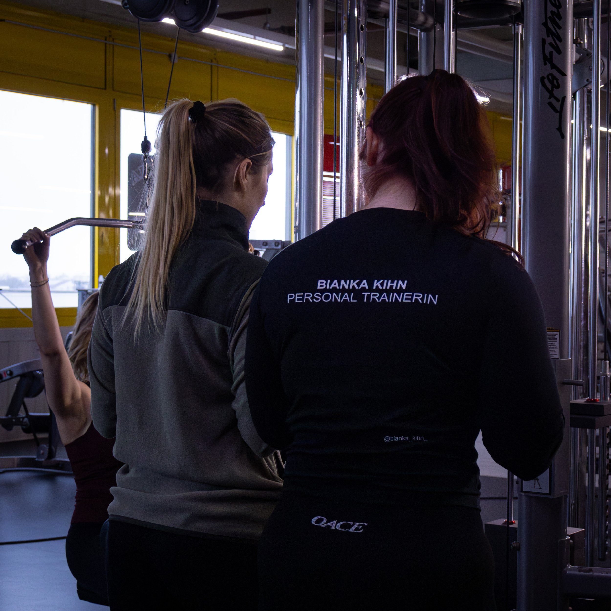 Drei Frauen in kurzer Kleidung im Fitnessstudio, eine davon trägt ein Shirt mit dem Namen 'Bianka Kihn', während sie an den Fitnessgeräten trainieren.