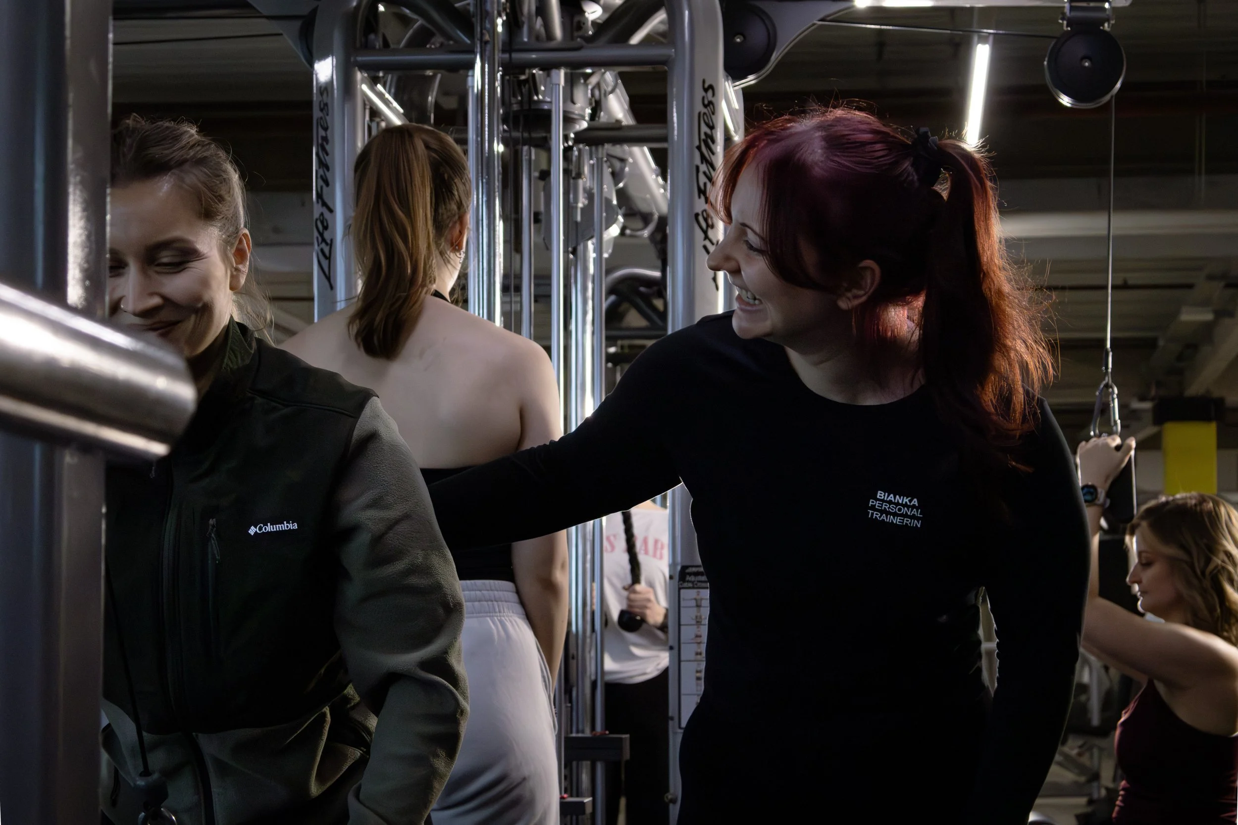 Menschen beim Krafttraining in einem Fitnessstudio, eine Trainerin hilft einer Frau beim Training, im Hintergrund eine Person mit langen Haaren, Spiegel und Fitnessgeräte.
