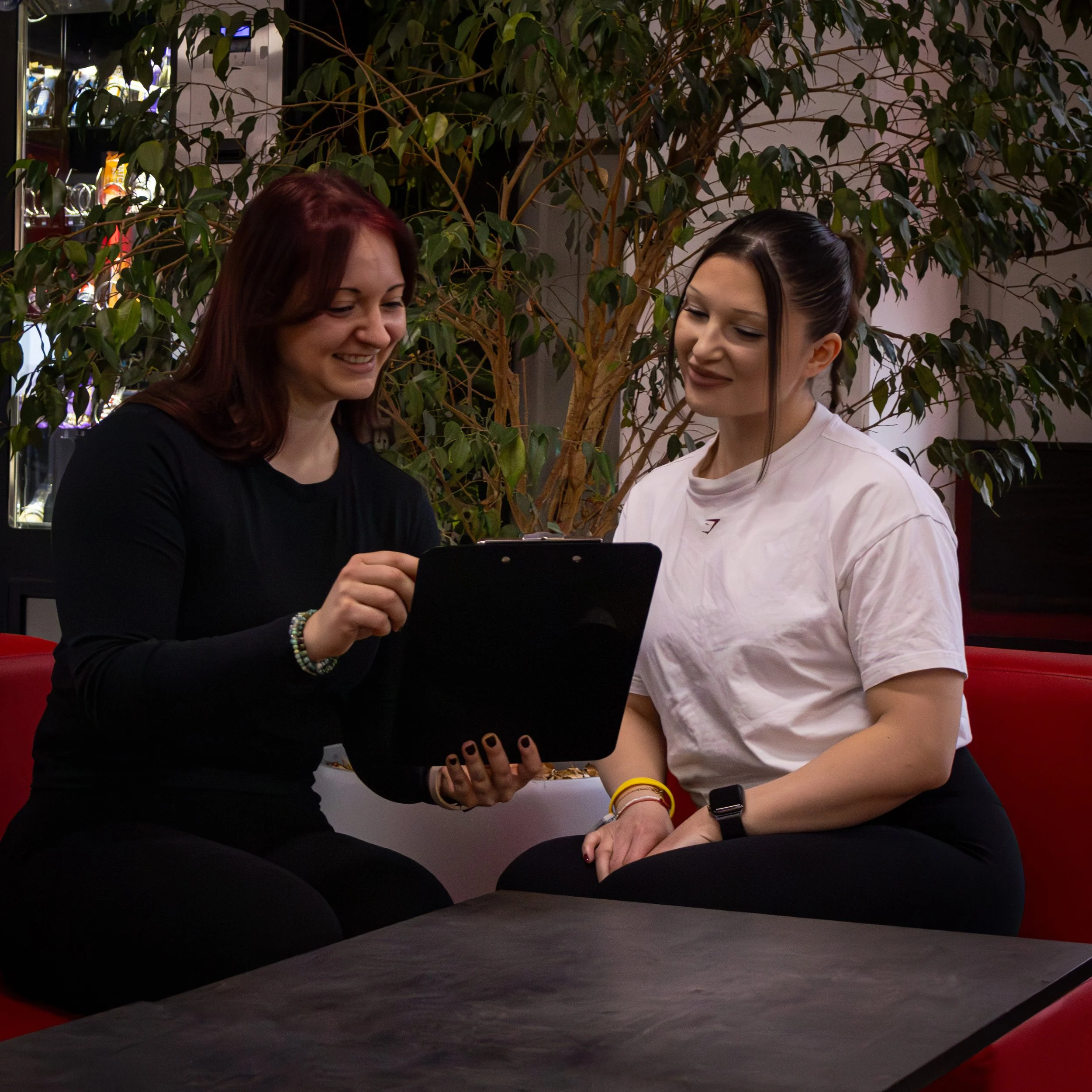 Zwei Frauen schauen zusammen auf ein Tablet, während sie in einem Saal sitzen, mit grünem Baum im Hintergrund.