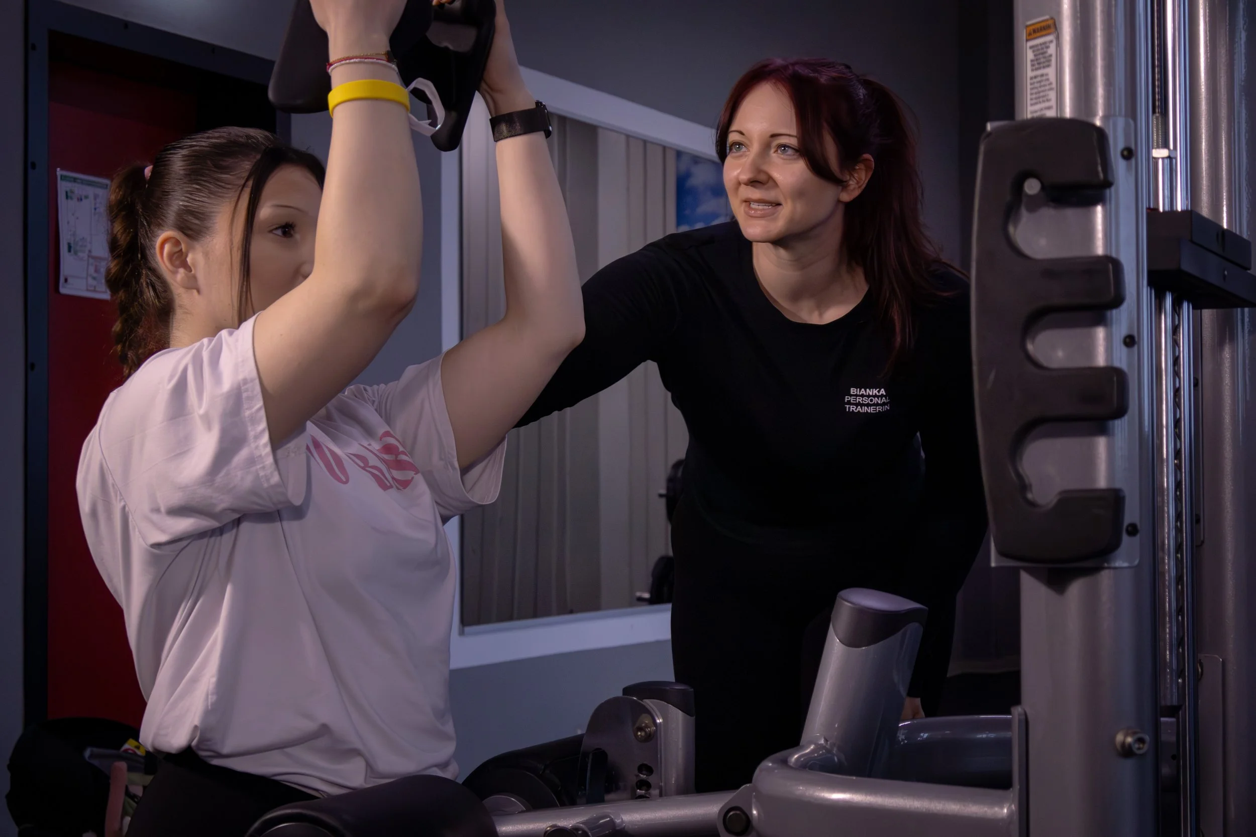 Eine Trainerin hilft einer jungen Frau bei Krafttraining an einer Maschine im Fitnessstudio.