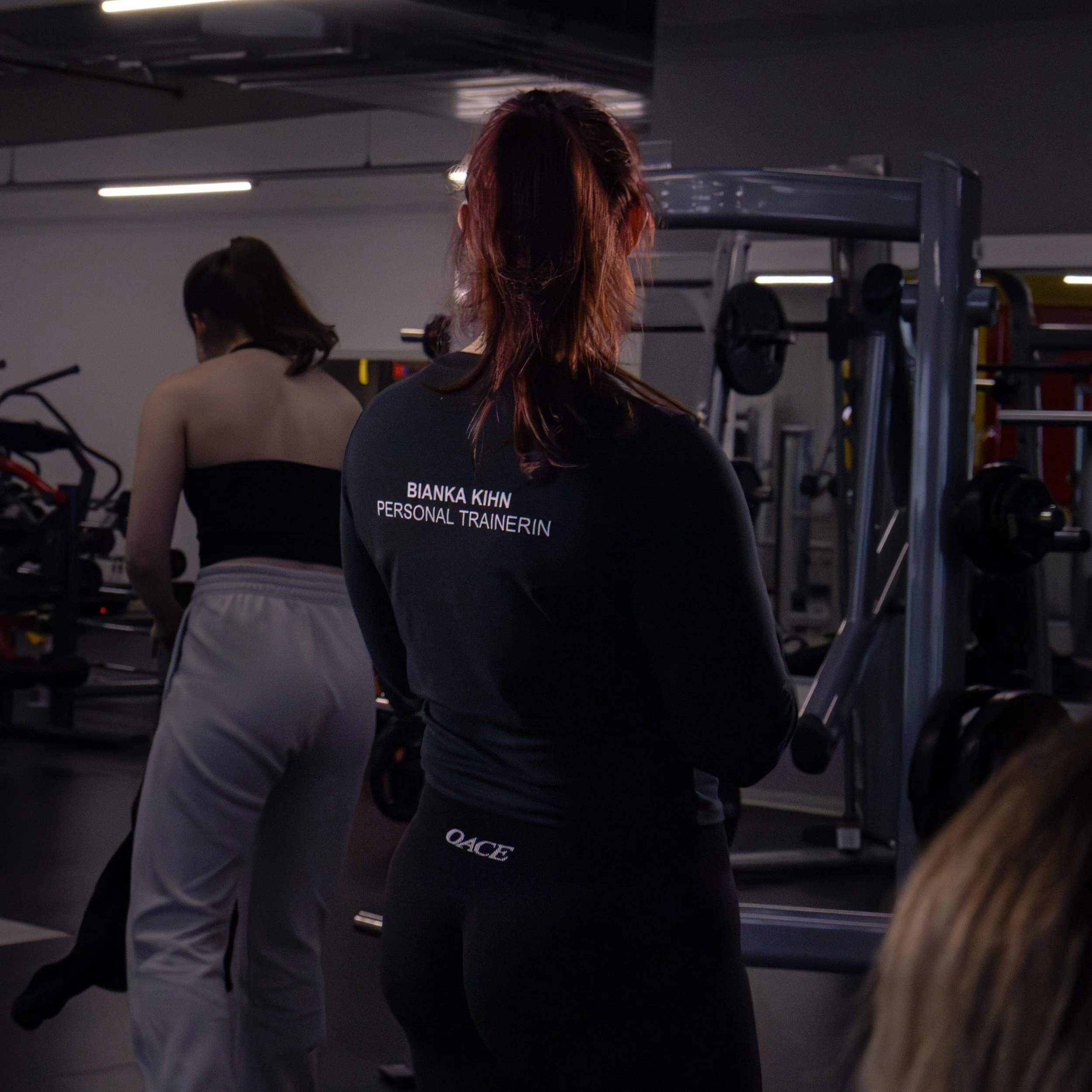 Zwei Frauen in einem Fitnessstudio, eine trägt ein schwarzes T-Shirt mit englischem Text, die andere trägt einen grauen Hoodie. Sie stehen vor Fitnessgeräten, im Hintergrund sind Gewichte und Sportgeräte sichtbar.