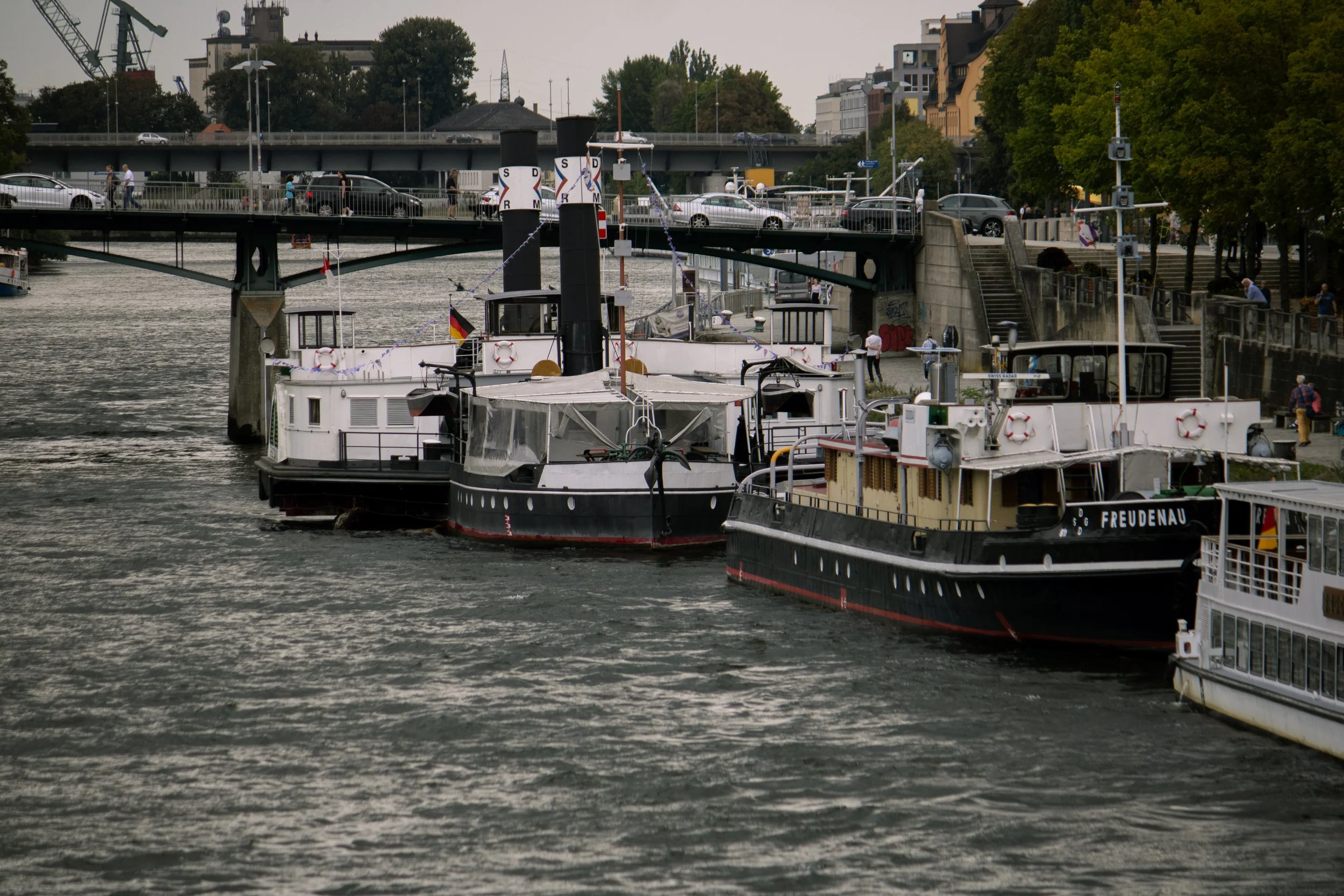 Der Fluss mit mehreren anlegenden Booten, über der Brücke fährt Verkehr, im Hintergrund Gebäude und Bäume.