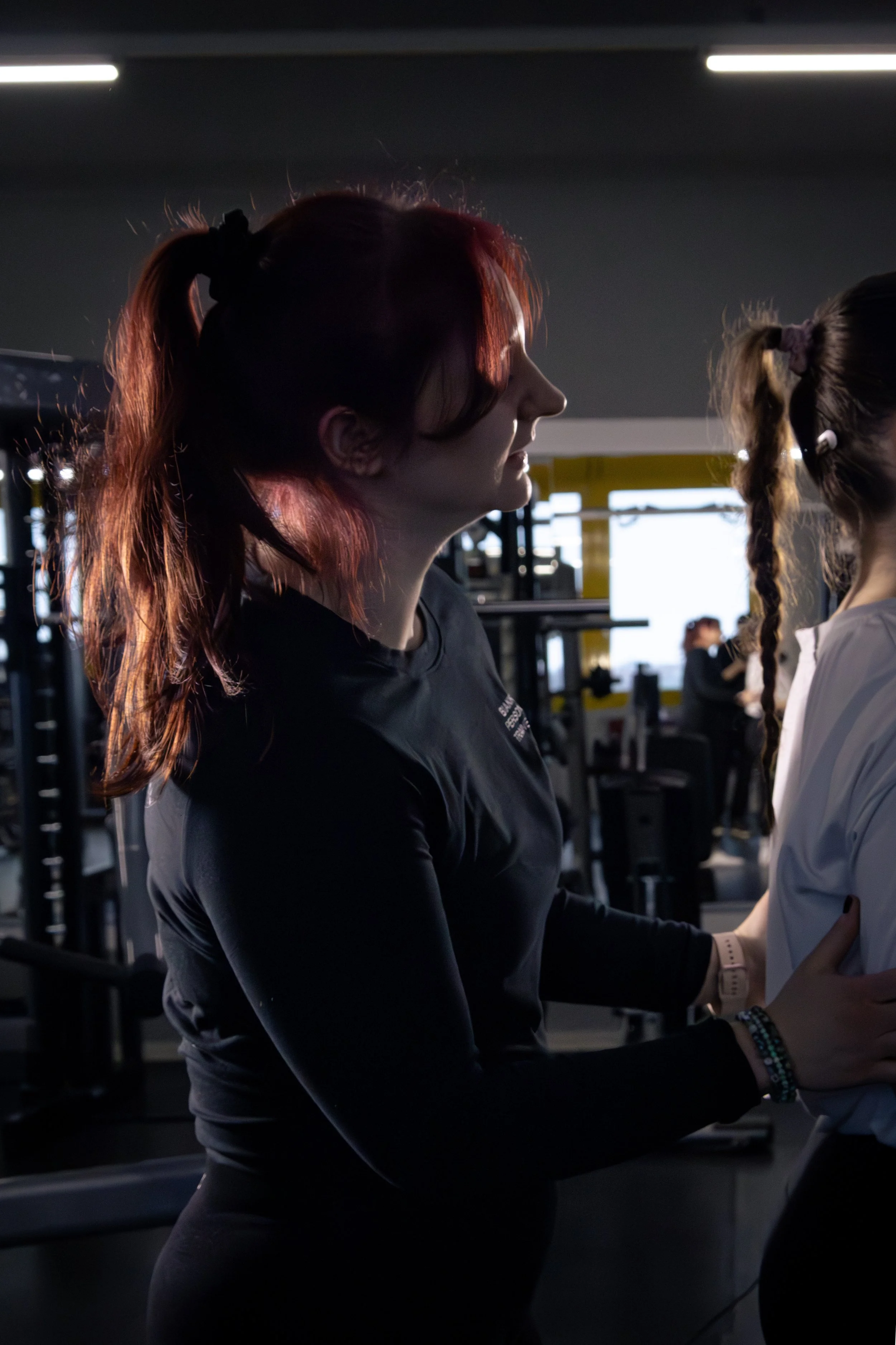 Zwei junge Frauen trainieren im Fitnessstudio, eine von ihnen trägt einen schwarzen Sport-BH und die andere ein graues Shirt, beide haben langes Haar im Pferdeschwanz, im Hintergrund sind Fitnessgeräte sichtbar.