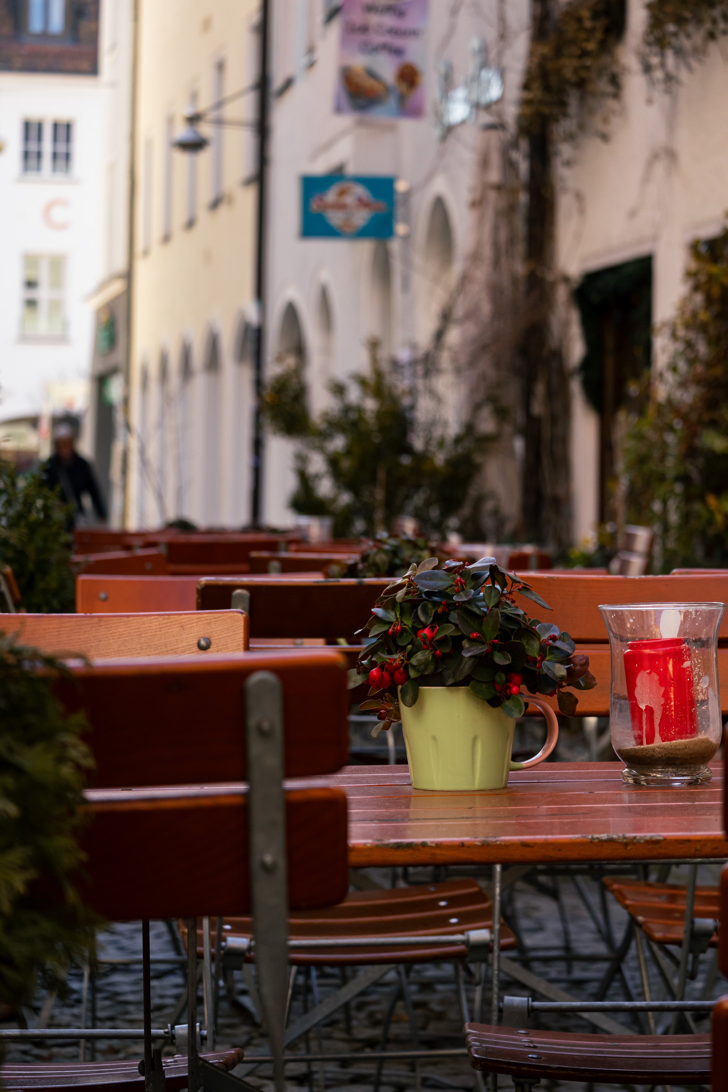 Ein Tisch im Freien mit Dekoration, darunter eine gelbe Tasse mit grünen Pflanzen und roten Beeren, sowie eine rote Kerze in einem Glas, umgeben von weiteren Stühlen und Tischen, im Hintergrund ein Spaziergänger und schlichte Gebäude.