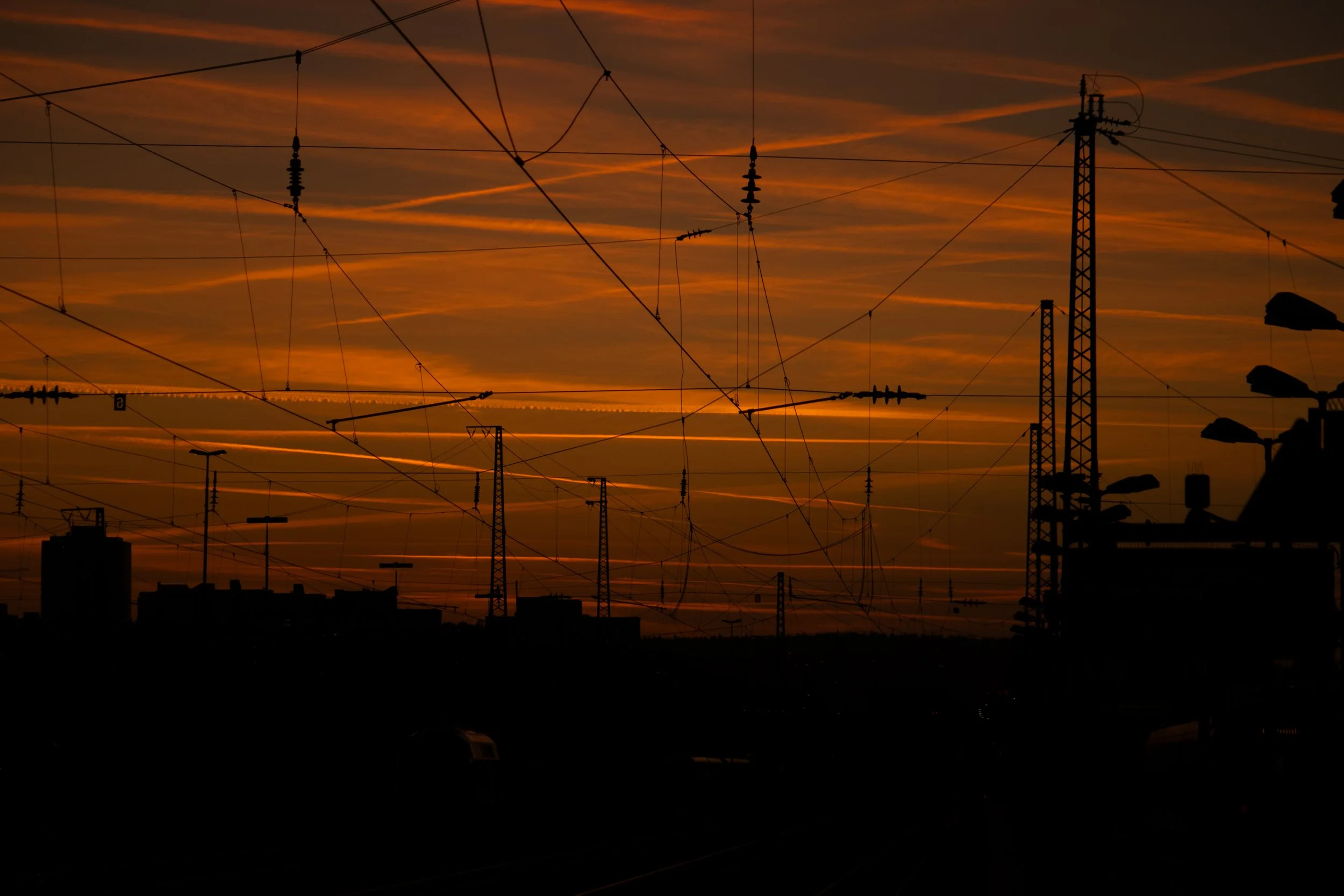 Silhouette von Eisenbahnleuchtmasten und Oberleitung vor einem orangefarbenen Sonnenuntergang.