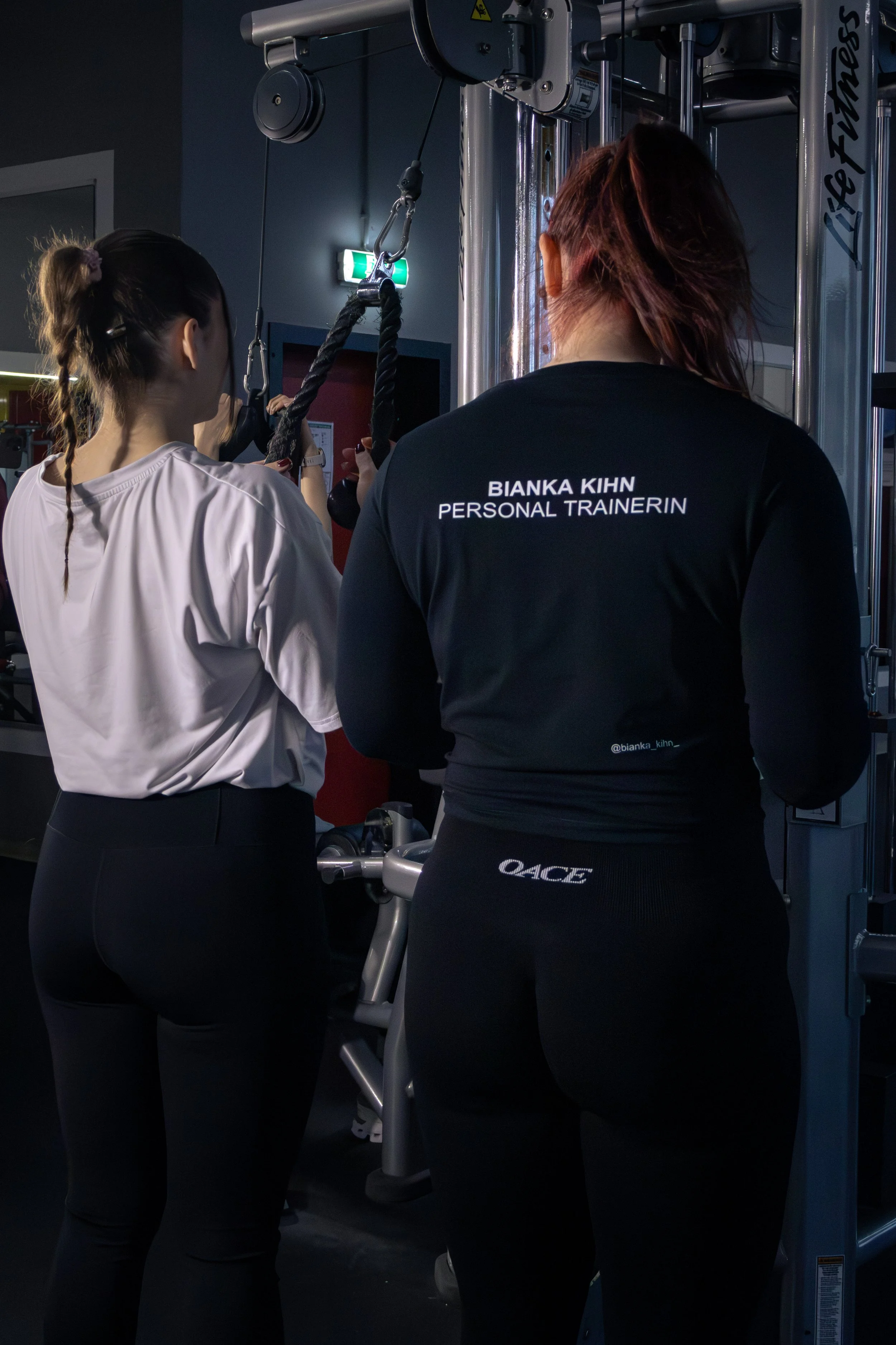 Zwei Frauen, eine mit dem Namen Bianka Kihn, trainieren an einem Kabelzug im Fitnessstudio, während sie von einer Trainerin angeleitet werden.