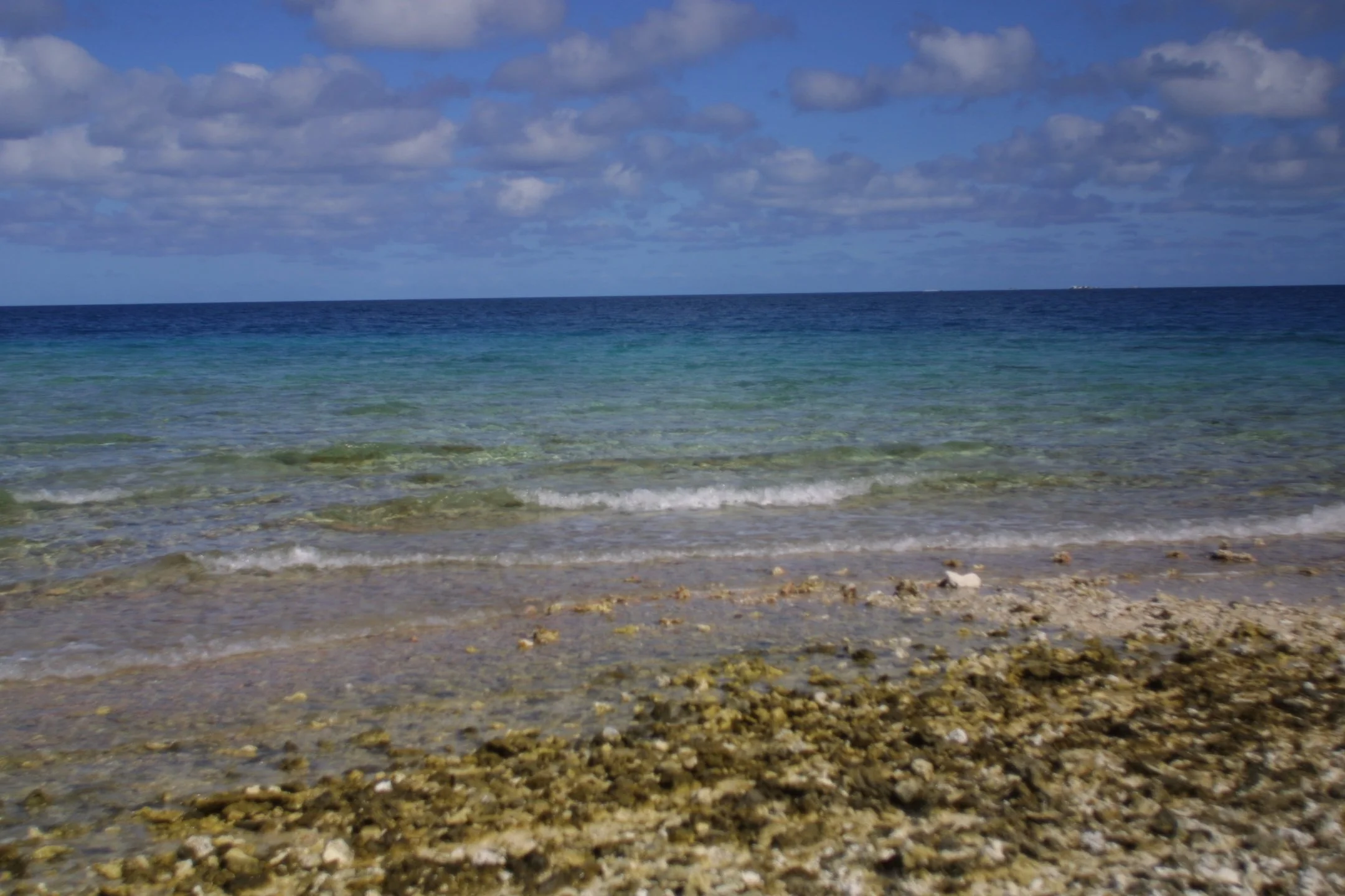 Photo of a calm ocean with gentle waves, a blue sky with scattered clouds, and a rocky sandy shoreline in the foreground.