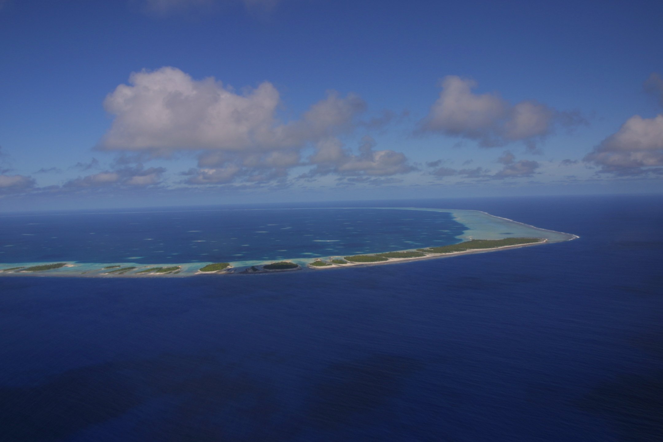 Aerial view of an island surrounded by blue ocean waters with a strip of land and a few small islands along the shoreline, under a partly cloudy sky.