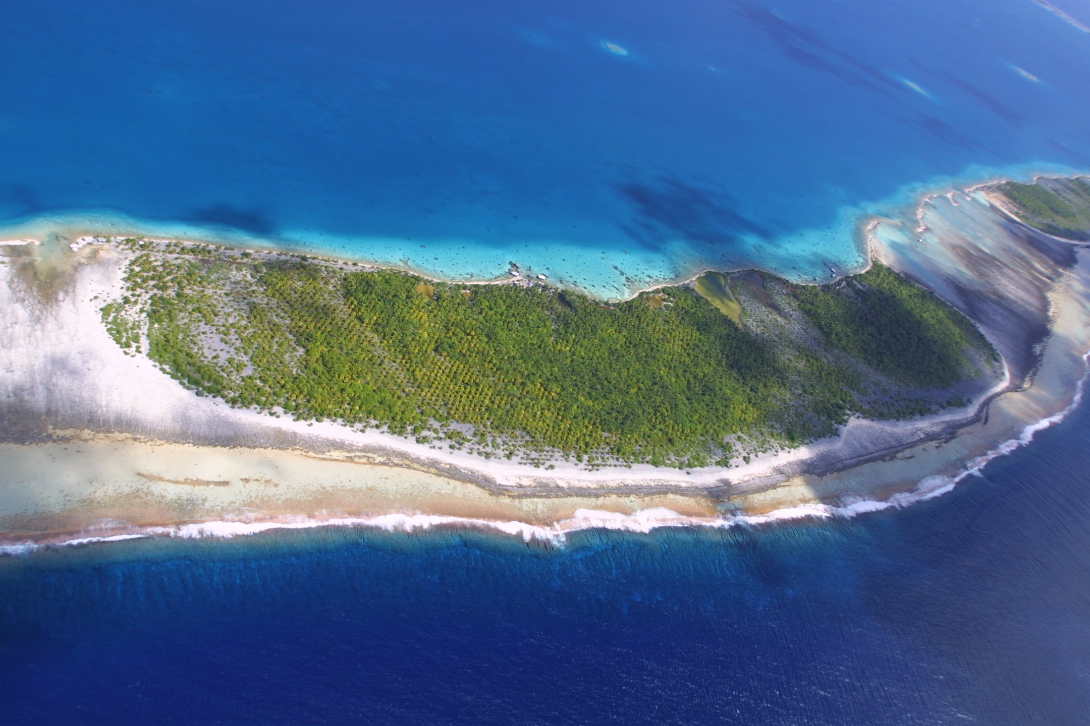 Aerial view of a small island with green vegetation, surrounded by blue ocean water with shades of turquoise near the shore. The island has a sandy beach and a narrow land bridge connecting to a larger area of land.