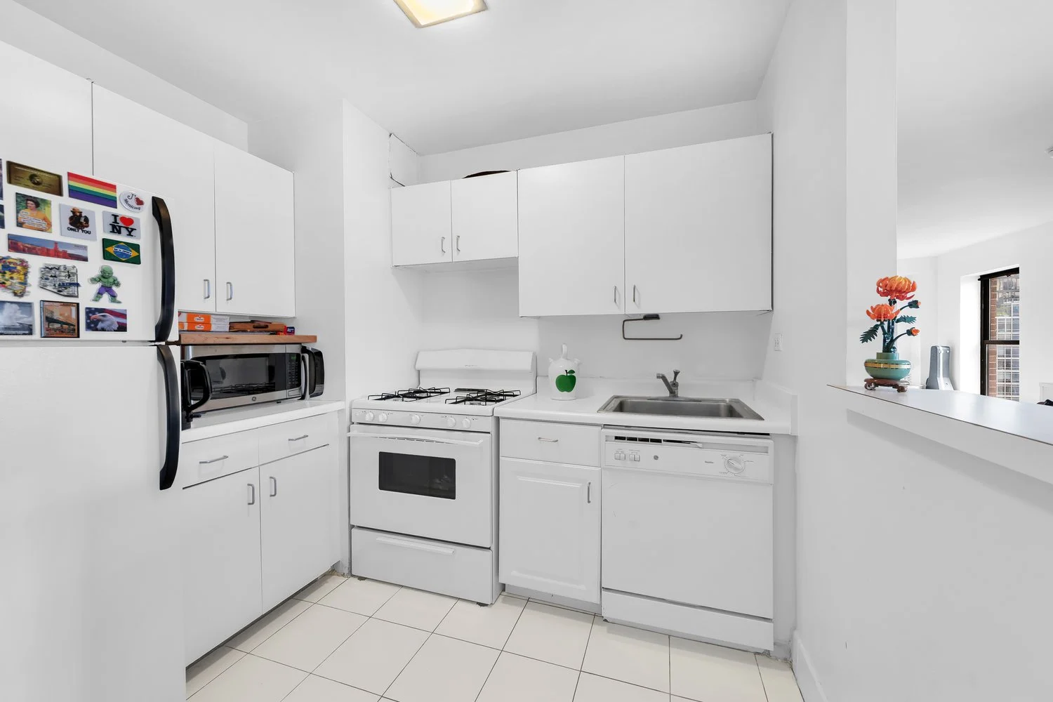 A white kitchen with a refrigerator covered in magnets, a microwave, a stove, a sink, and a dishwasher. There is a green apple paper towel holder on the counter and a vase with orange flowers on the windowsill.