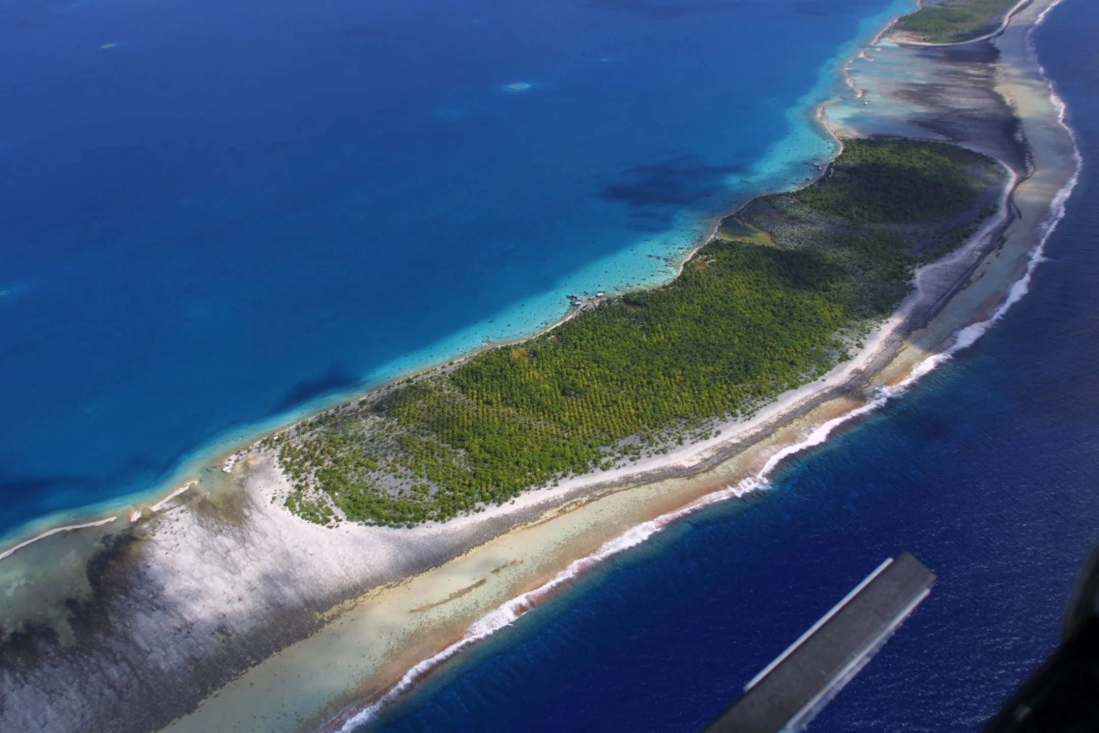 Aerial view of a tropical island with lush green vegetation, surrounded by turquoise and deep blue ocean waters, with a peninsula extending into the sea.