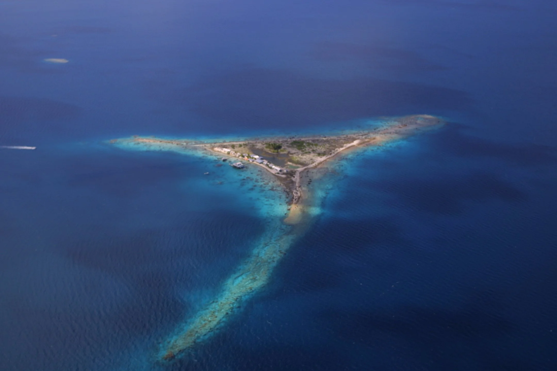 An aerial view of a small island surrounded by blue ocean waters, with a few structures and trees visible on the island.