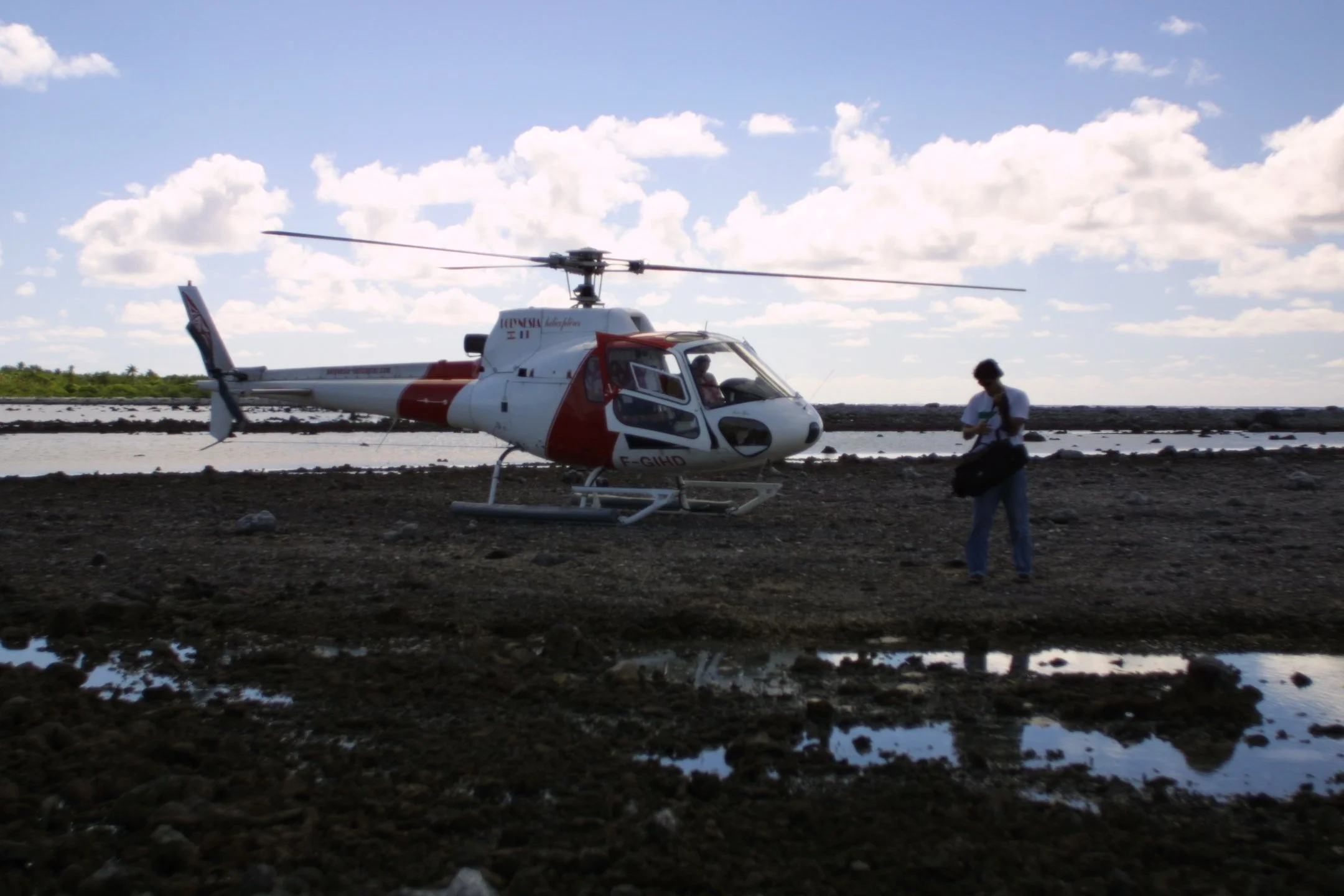 A helicopter parked on rocky ground near a body of water under a partly cloudy sky, with a person walking nearby carrying a bag.