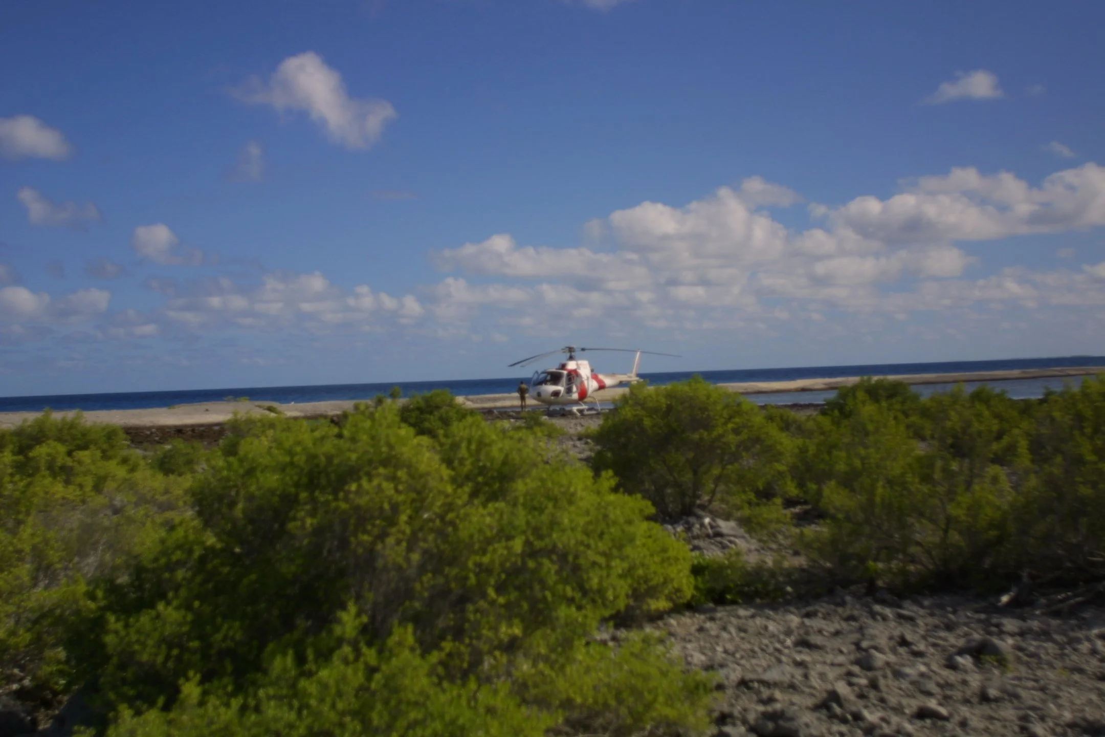 A white and red helicopter on the ground near the shoreline with blue sky and clouds in the background.