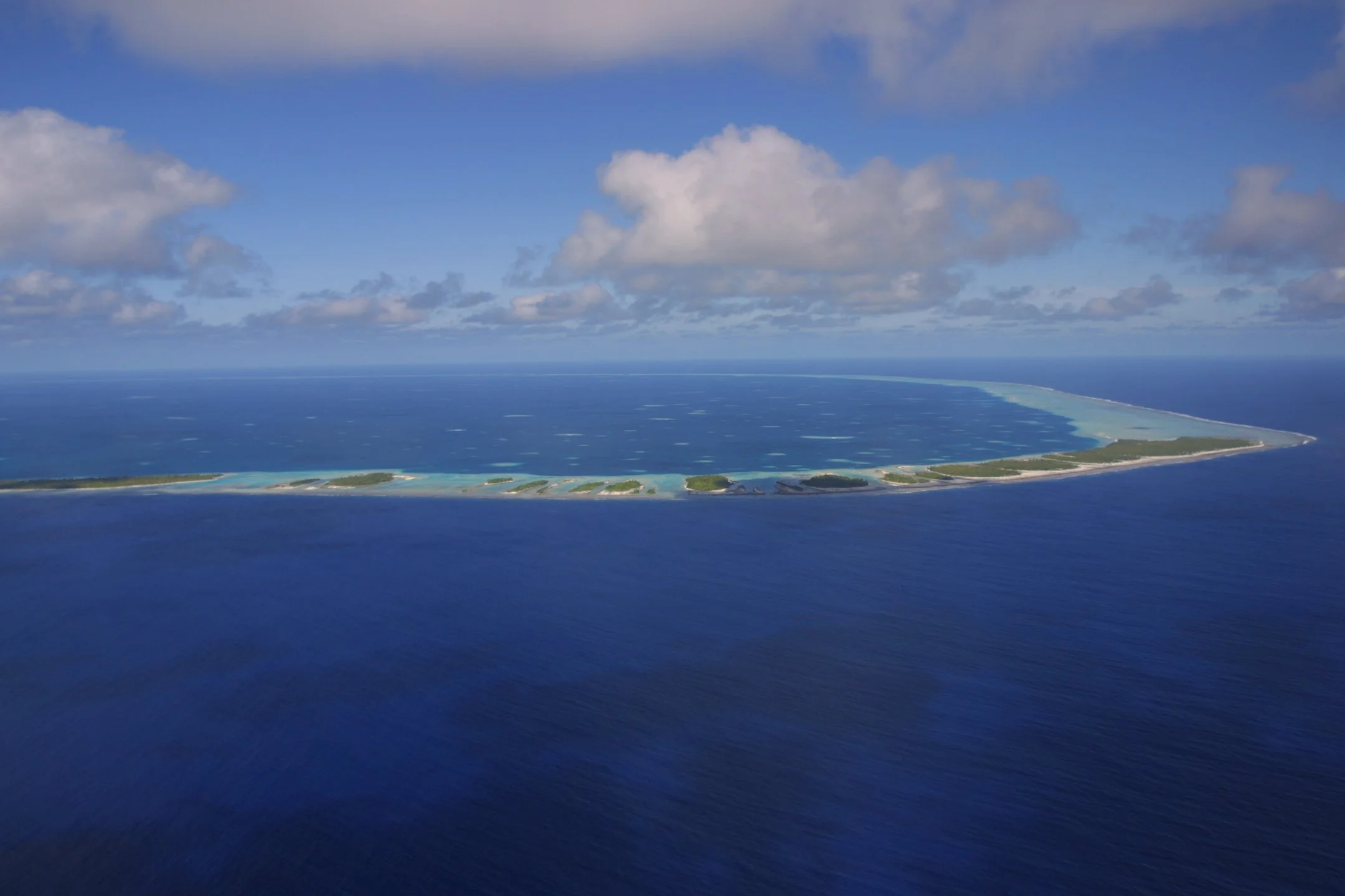 An aerial view of a long, curved island surrounded by blue ocean water with some greenery and sandy beaches visible on the island, under a partly cloudy sky.