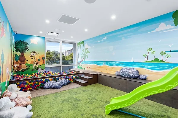 Indoor play area with colorful murals of a jungle on one wall and a beach scene on another, featuring a small green slide, stuffed animals, and a ball pit with multicolored balls at 1399 Park Avenue luxury condo upper east side new york