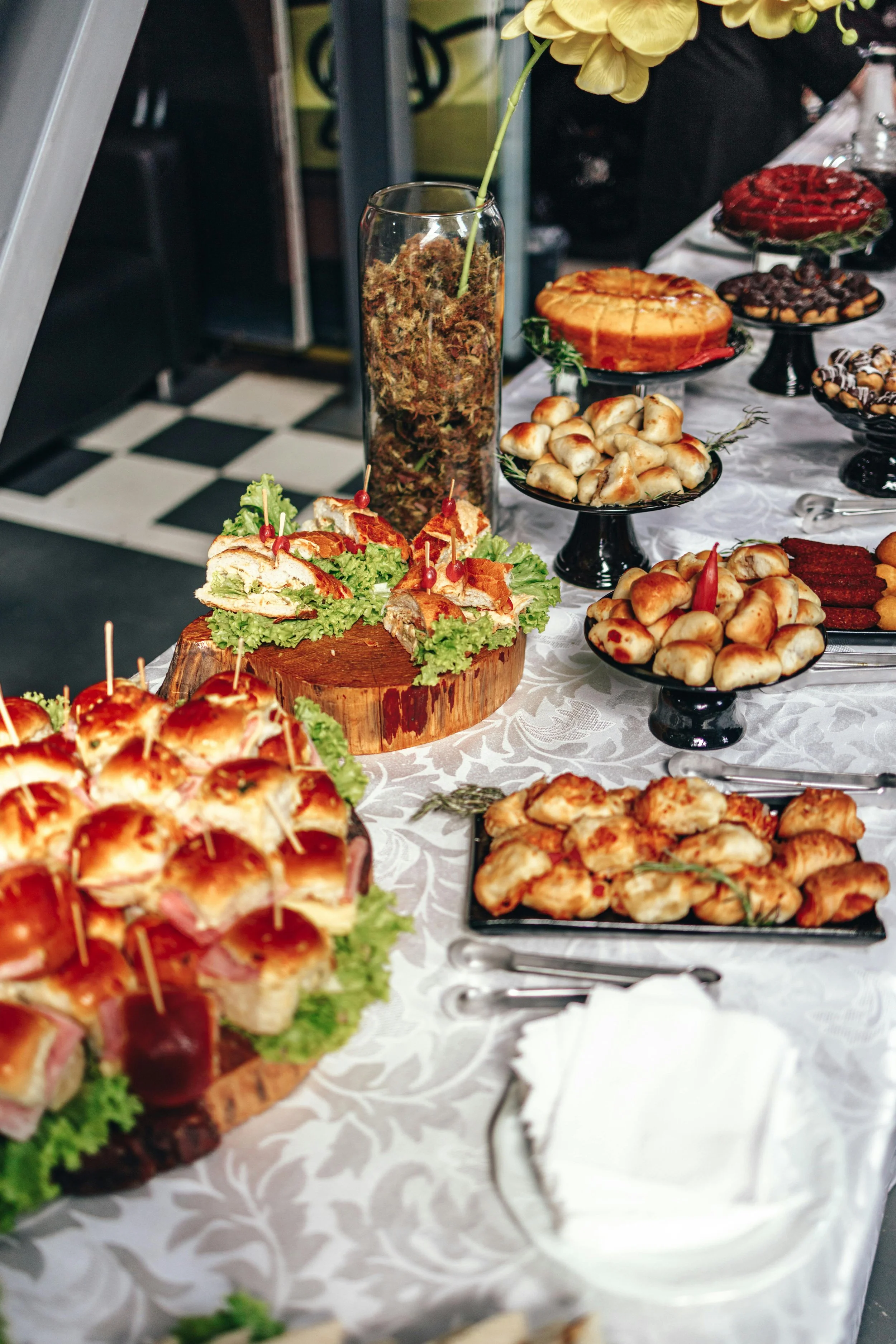 A buffet table with various foods including mini sandwiches with lettuce and bacon, small pastries, cheesecakes, and candies on black stands, decorated with yellow flowers, on a white tablecloth.