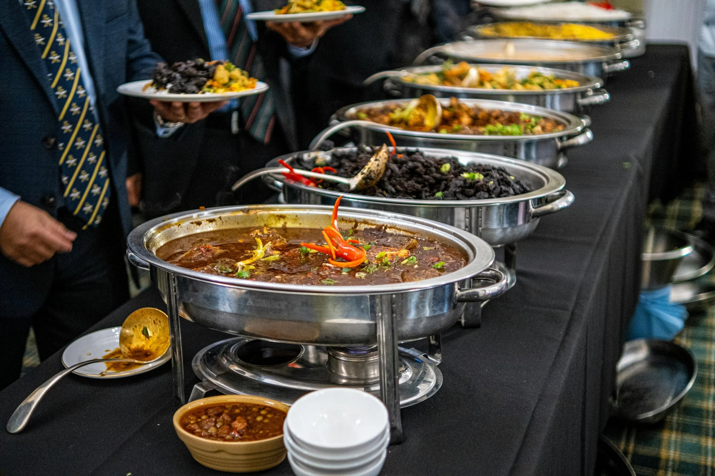 A variety of chafing dishes filled with different types of hot foods at a buffet, with people serving themselves.