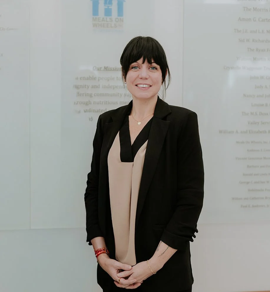 A woman with black hair and blue eyes, wearing a black blazer over a beige top, standing in front of a white wall with some blurred text and logos.