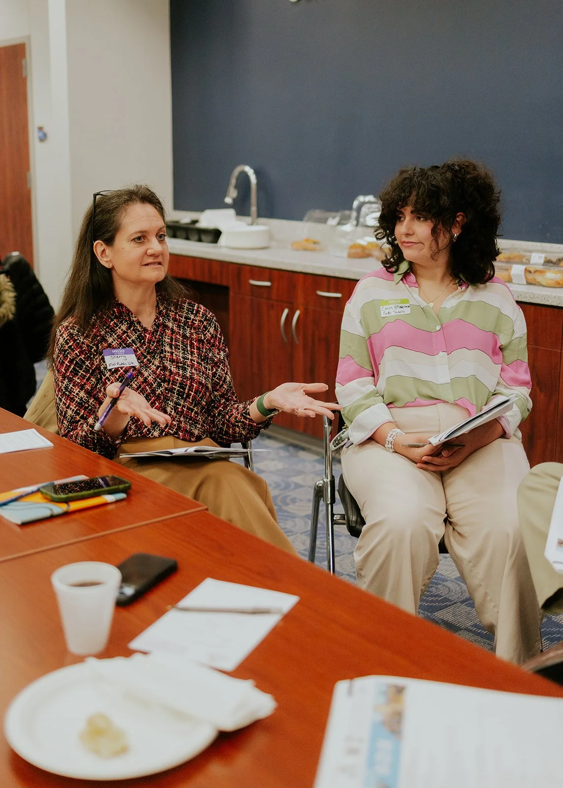 Two women sitting at a conference table during a meeting, engaging in conversation. One woman is gesturing with her hands, holding a pen, and the other woman is listening, holding a notepad. There are papers, a coffee cup, and a smartphone on the table. The background shows a kitchen counter with a sink and some snacks.