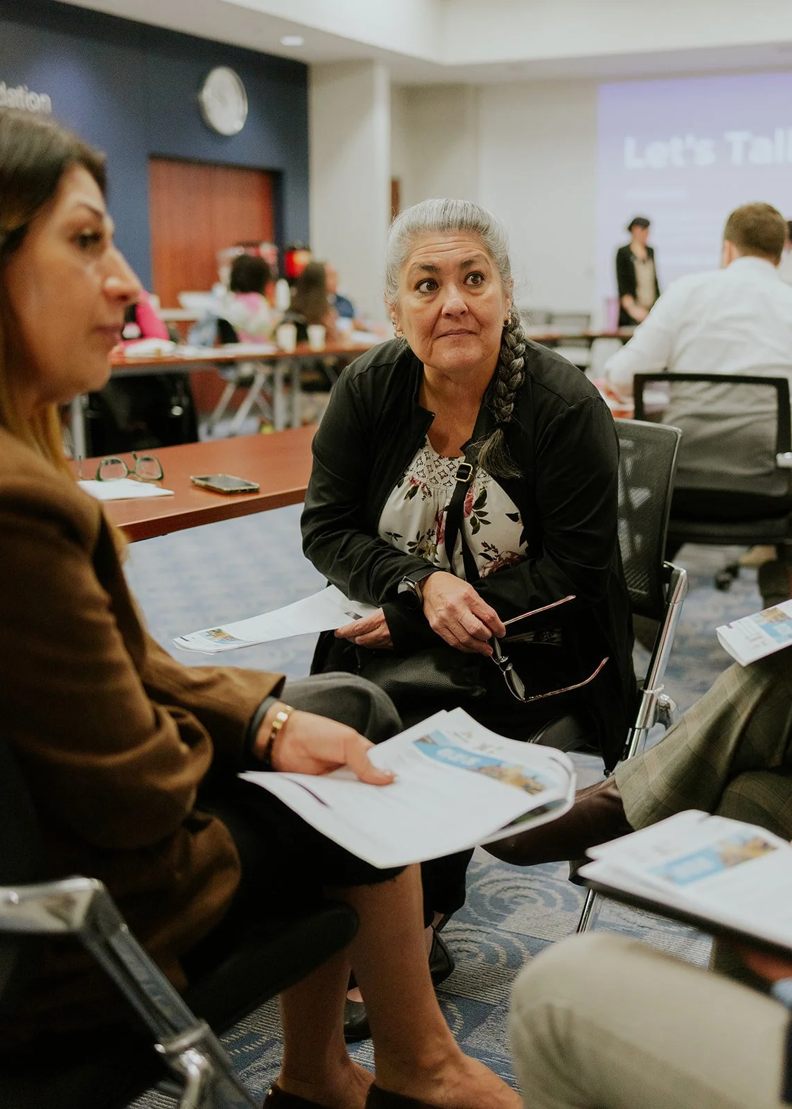 A woman with gray hair in a braid, wearing a black jacket and floral blouse, sitting at a table during a conference or meeting in a large room.