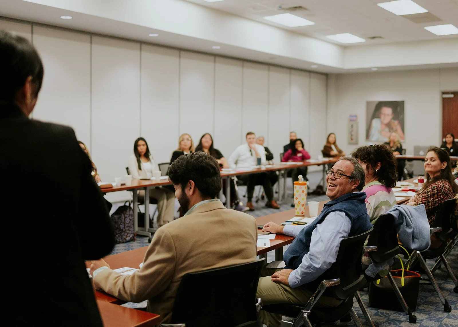 A group of diverse people seated around a U-shaped conference table, engaging in a meeting or workshop inside a well-lit room with gray walls. Several participants are smiling, and one individual appears to be speaking or presenting.