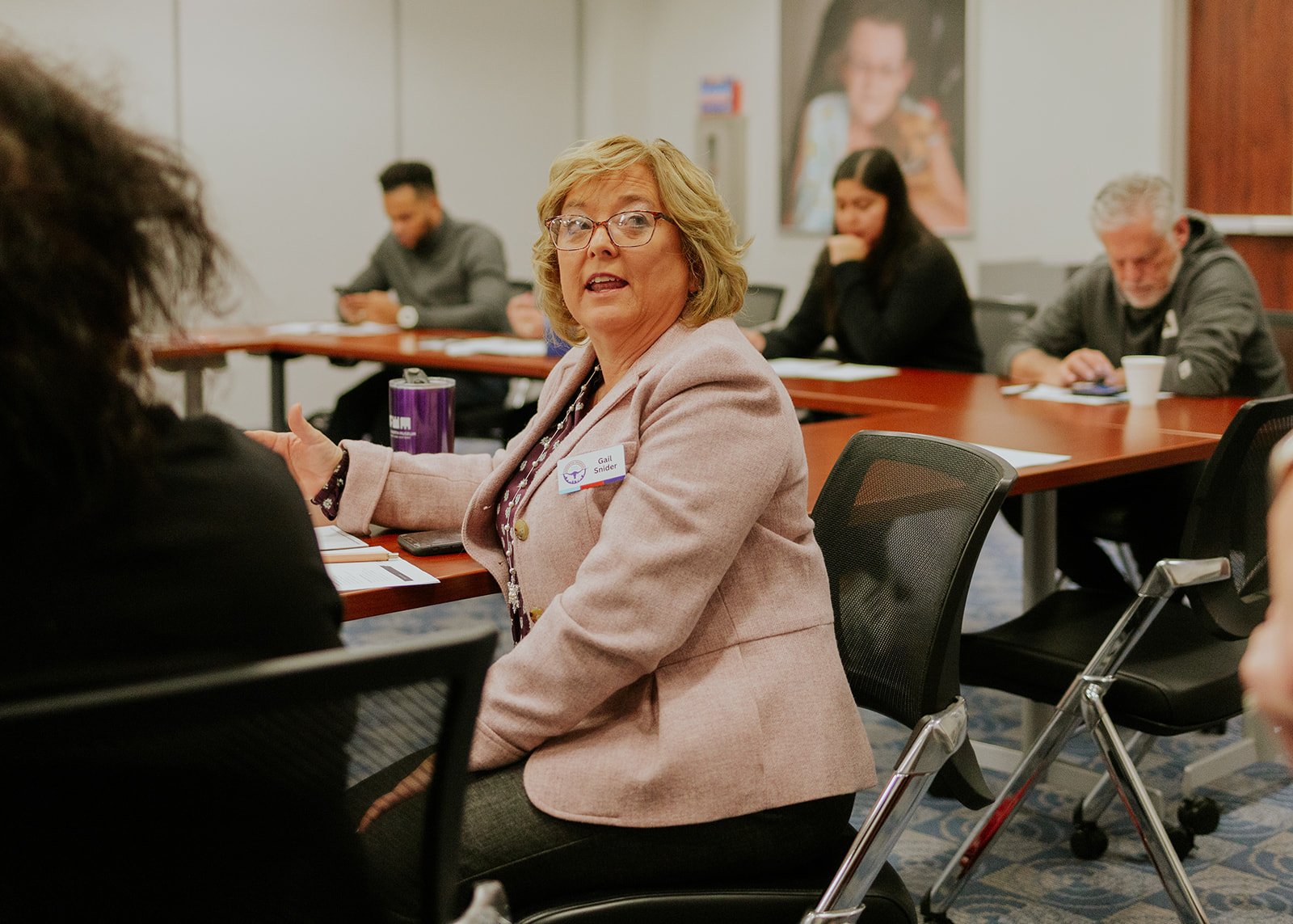 Woman with glasses and a pink blazer speaking at a conference table, with people listening and taking notes in the background