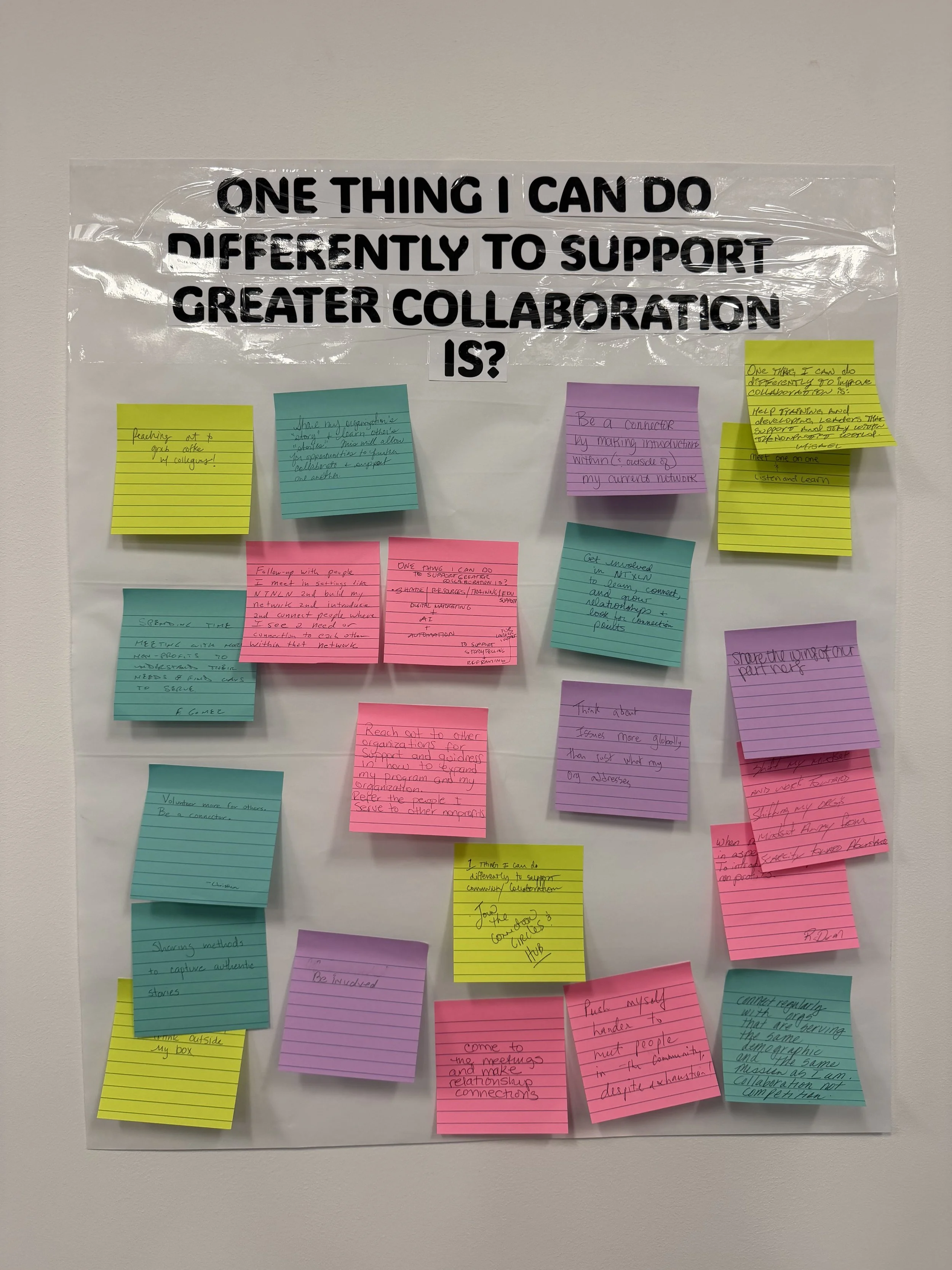 A wall with multiple colorful sticky notes containing handwritten text, centered around a large transparent sign with bold black text that reads, 'ONE THING I CAN DO DIFFERENTLY TO SUPPORT GREATER COLLABORATION IS?'.