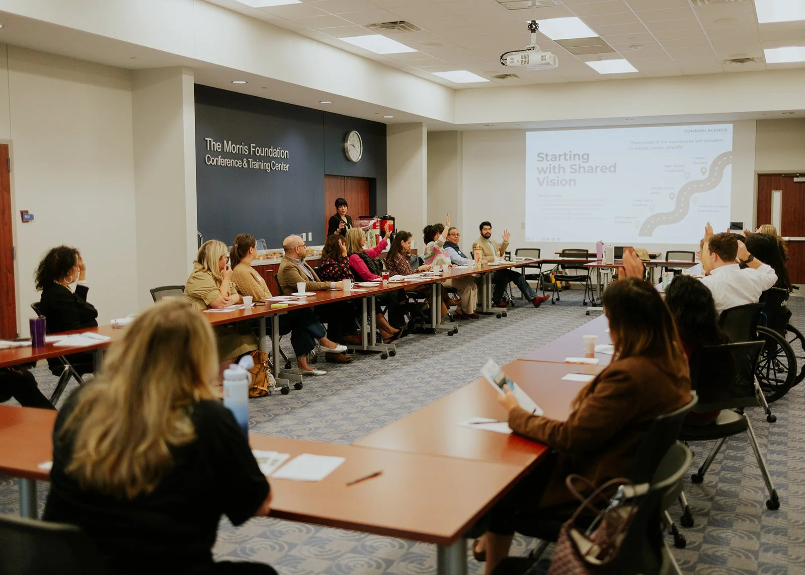 People attending a conference or training session in a large meeting room, with some participants raising their hands during a discussion, and a presenter standing at the front near a screen with a presentation slide.