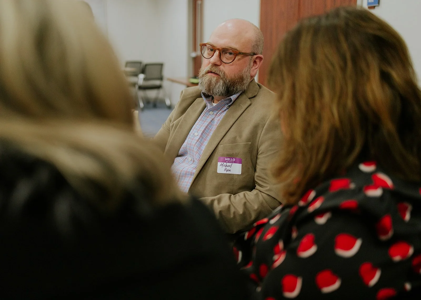 A man with glasses, a beard, and a bald head wearing a tan blazer and a name tag labeled "Michael Ryon" is sitting at a discussion table with two women, one with blonde hair and the other with brown hair and a black top with red and white patterns, in a conference room.