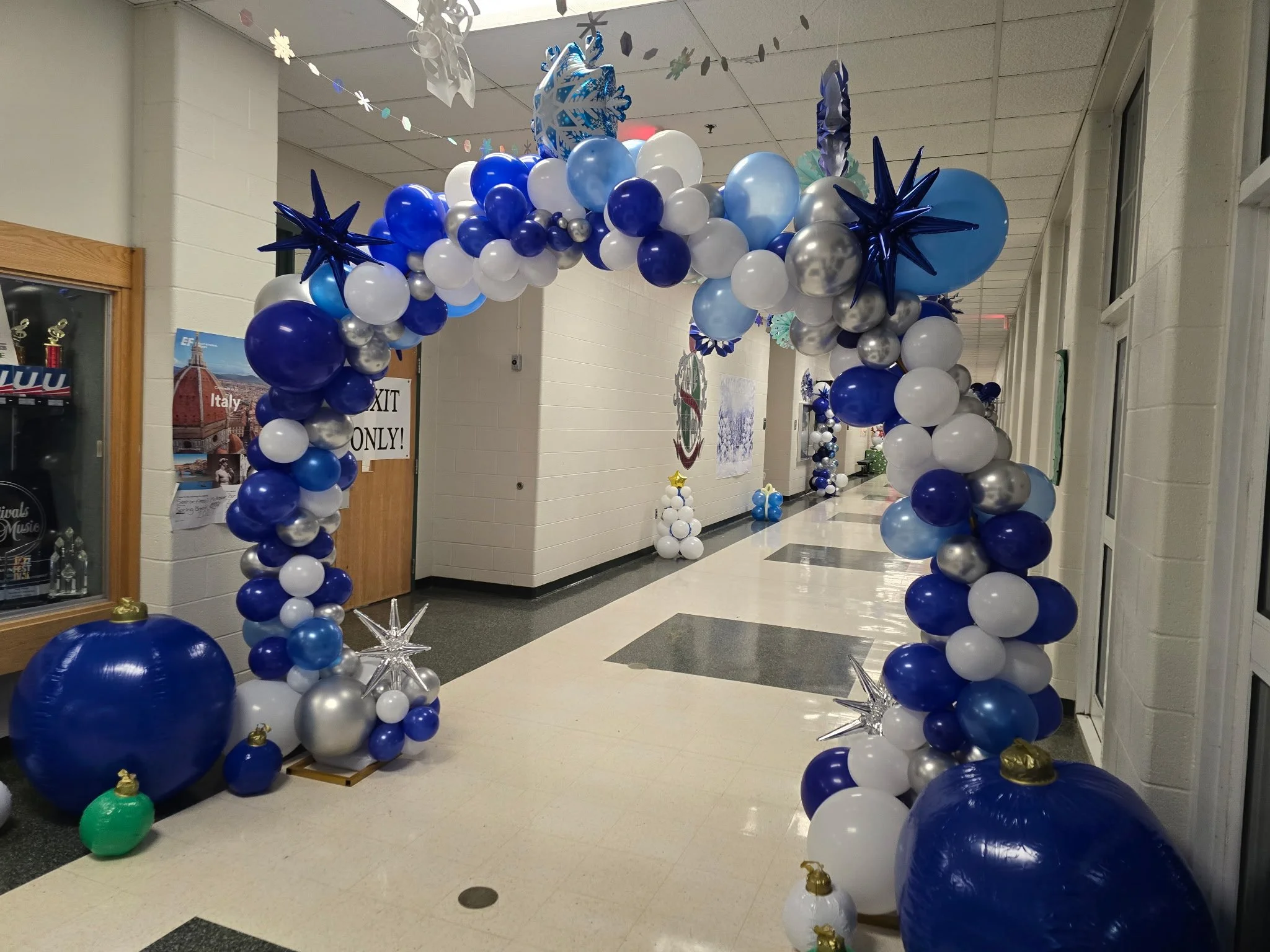 Hallway decorated with a large balloon arch and balloon clusters in blue, white, silver, and gold for celebration.