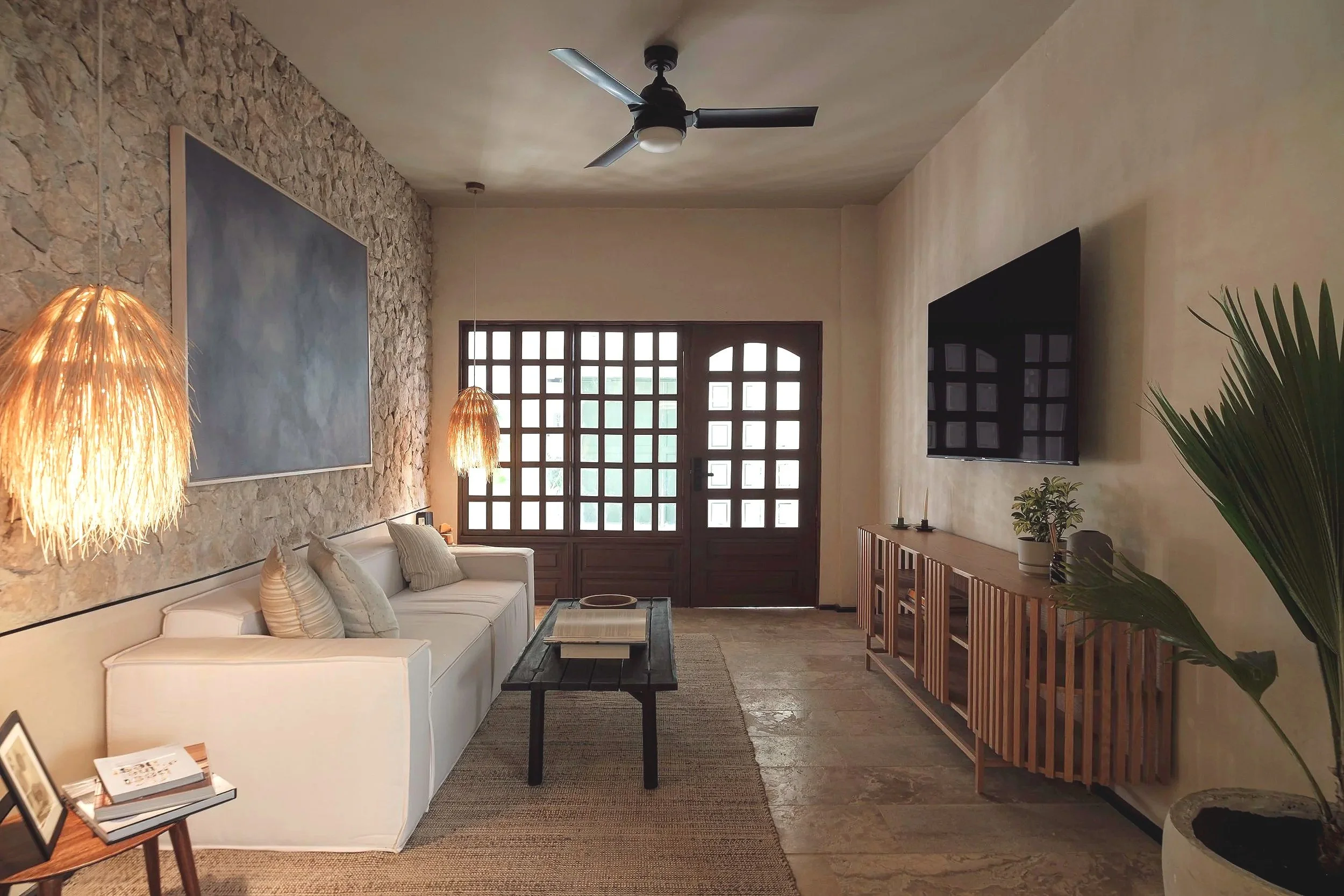 Living room with a white sofa, lamps, a large window with wooden grid pattern, a flat-screen TV on a light-colored wall, and decorative plants.