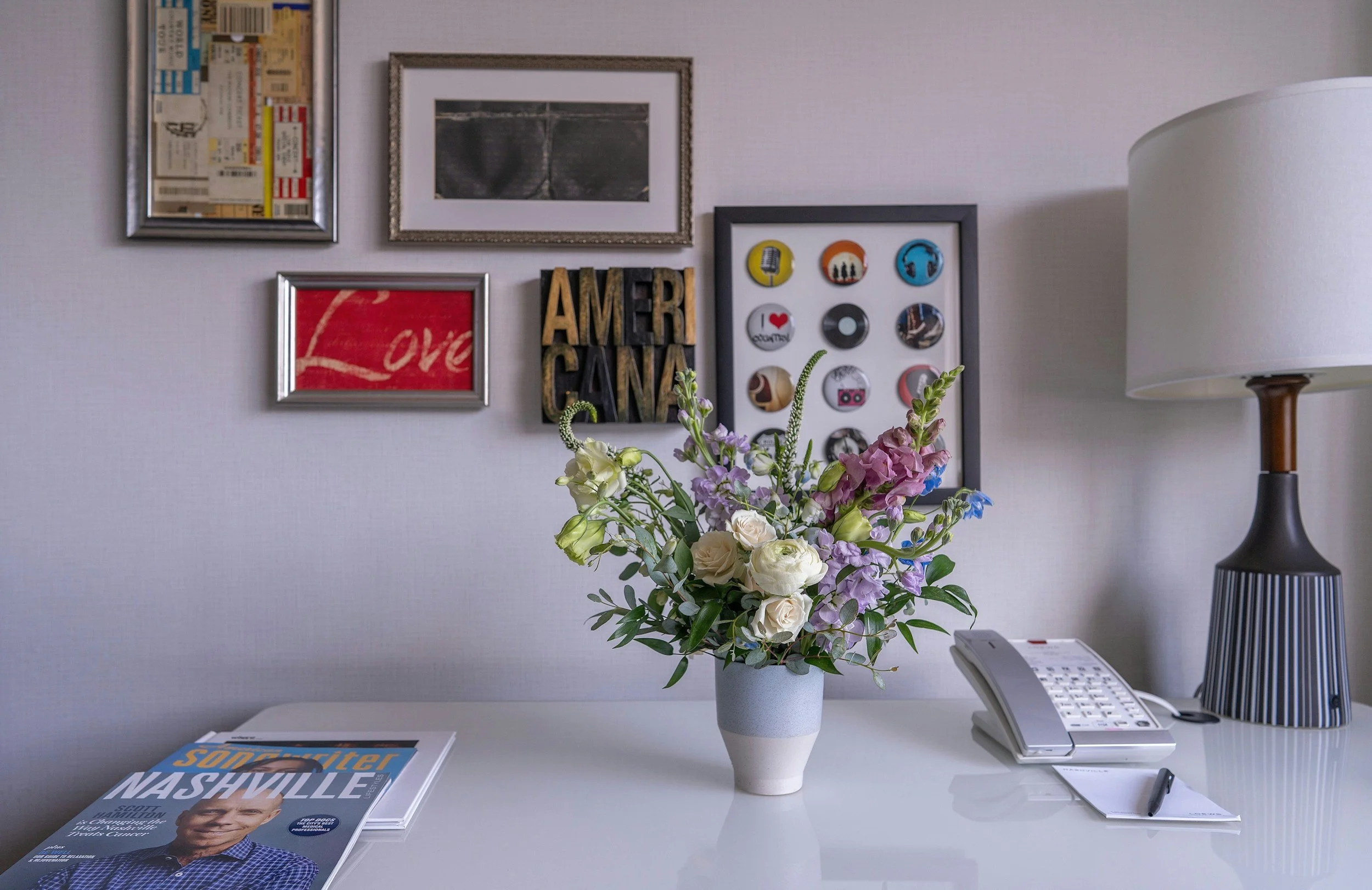 A white desk with a vase of mixed flowers, a magazine titled Nashville Scott, a telephone, and a pen. Behind the desk is a wall decorated with framed pictures and wall art, including a collage of buttons and a framed photograph. There is a lamp with a white shade and wooden base on the right side of the image.