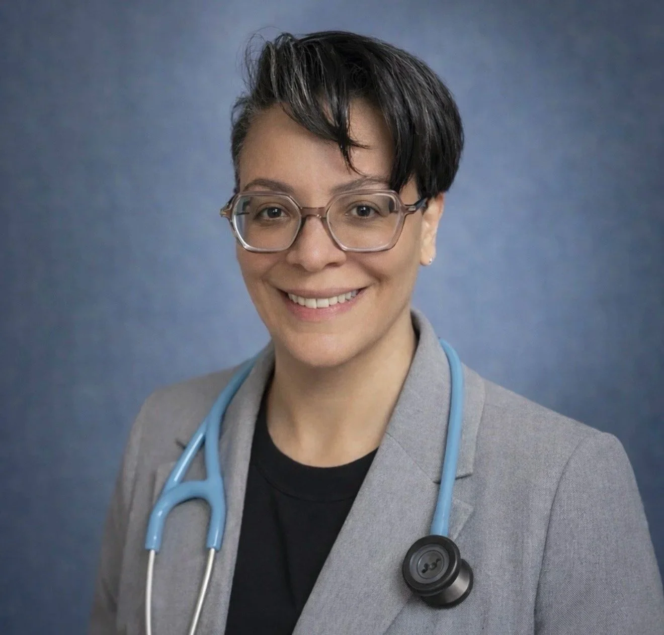 Professional woman doctor or healthcare provider with short dark hair, glasses, wearing a gray blazer and black shirt, with a blue stethoscope around her neck, smiling against a blue background.