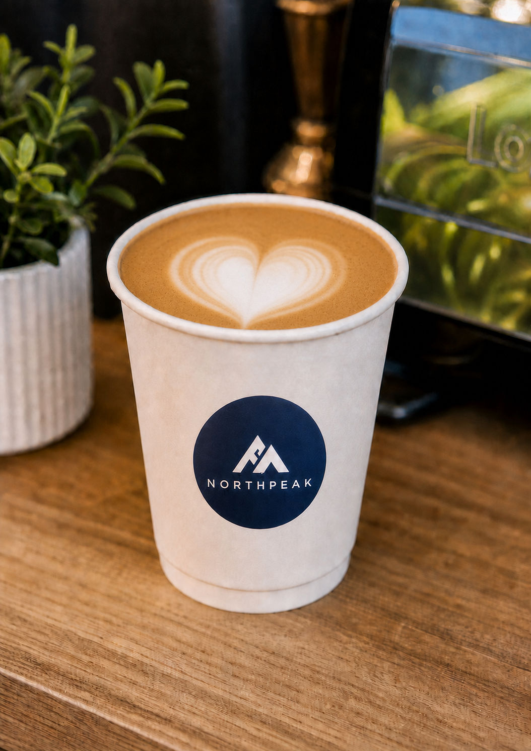 A paper cup of coffee with a heart-shaped latte art on top, placed on a wooden surface, with a small potted plant and coffee machine in the background.