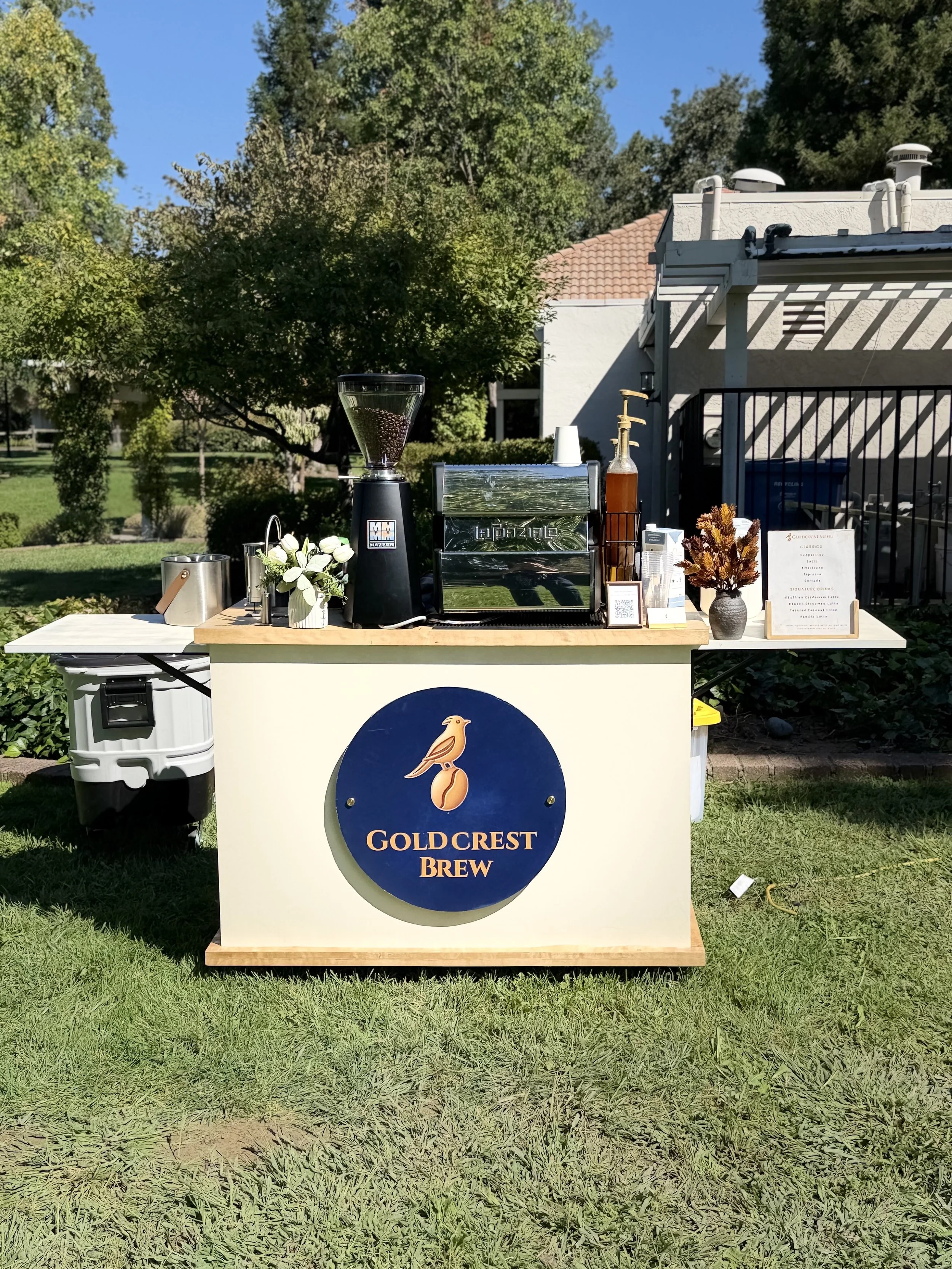 A beverage station labeled "Goldcrest Brew" featuring a coffee grinder, a syrup pump, flowers, and a menu, set outdoors on a grassy area with trees and a building in the background.