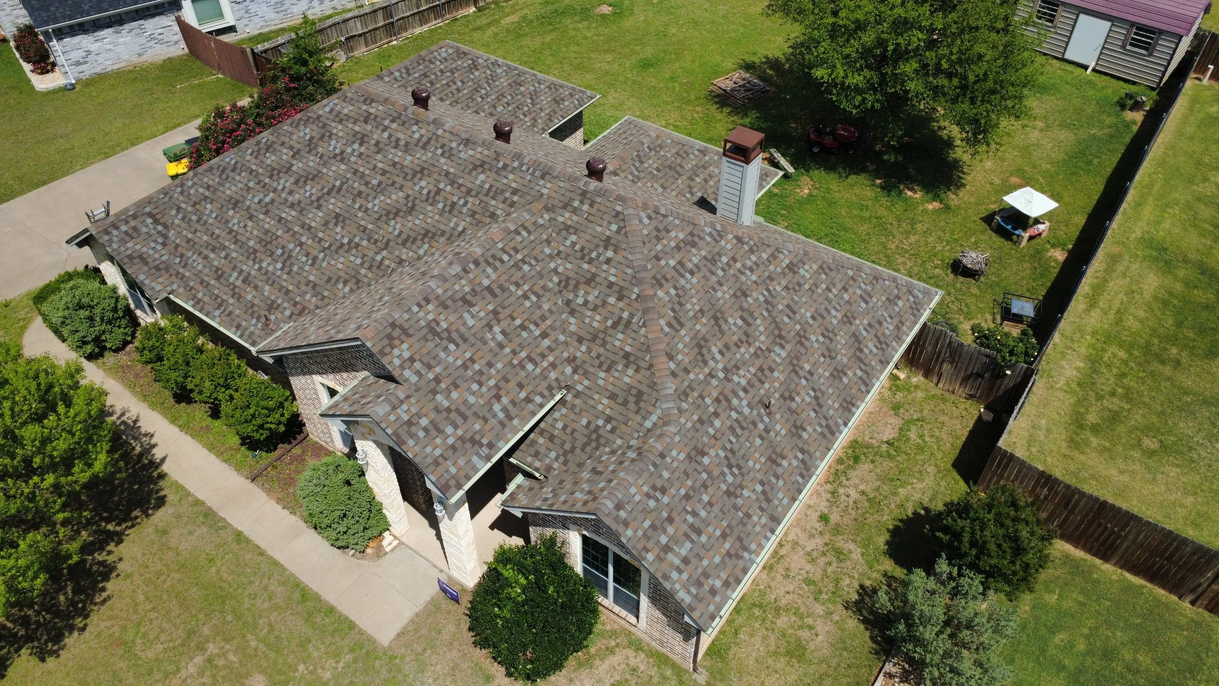 Aerial view of a house with a multi-gabled roof covered with brown shingles, surrounded by green lawns, trees, and neighboring houses and fences.