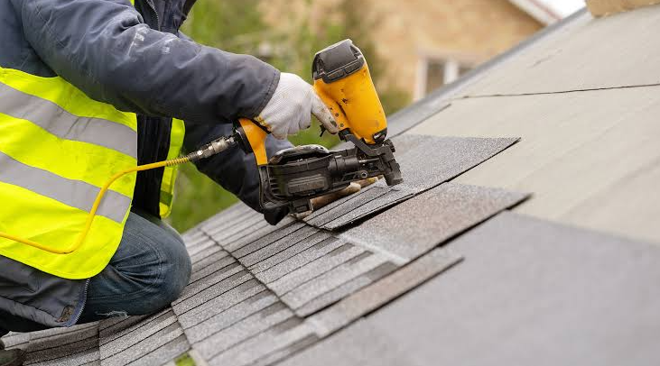 A worker installing shingles on a roof using a nail gun. The worker is wearing a high-visibility yellow vest and gloves.