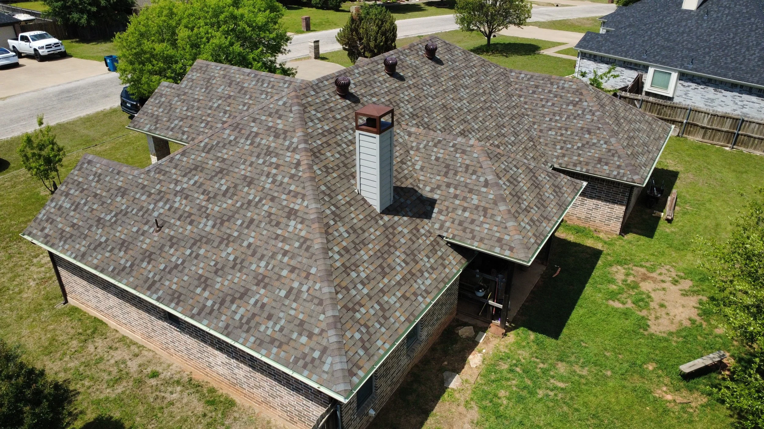 Aerial view of a house with a multicolored shingle roof, brick exterior walls, a chimney, surrounding green lawn, trees, and neighboring houses and vehicles.