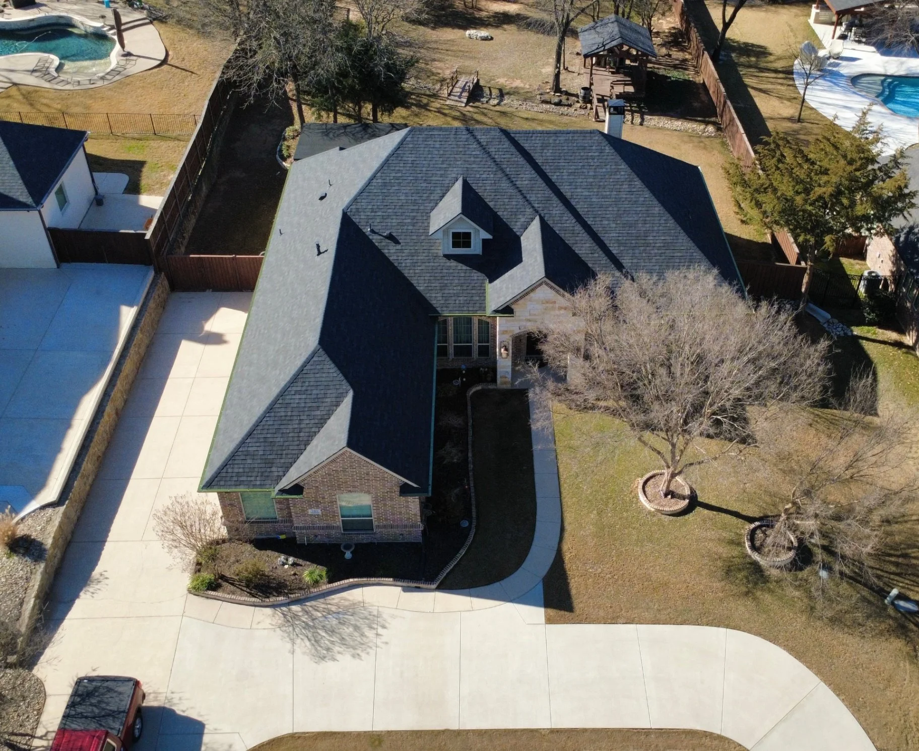 An aerial view of a residential backyard showing a house with a gray roof, fenced-in yard, two leafless trees, a curved concrete walkway, swimming pools, and outdoor structures.